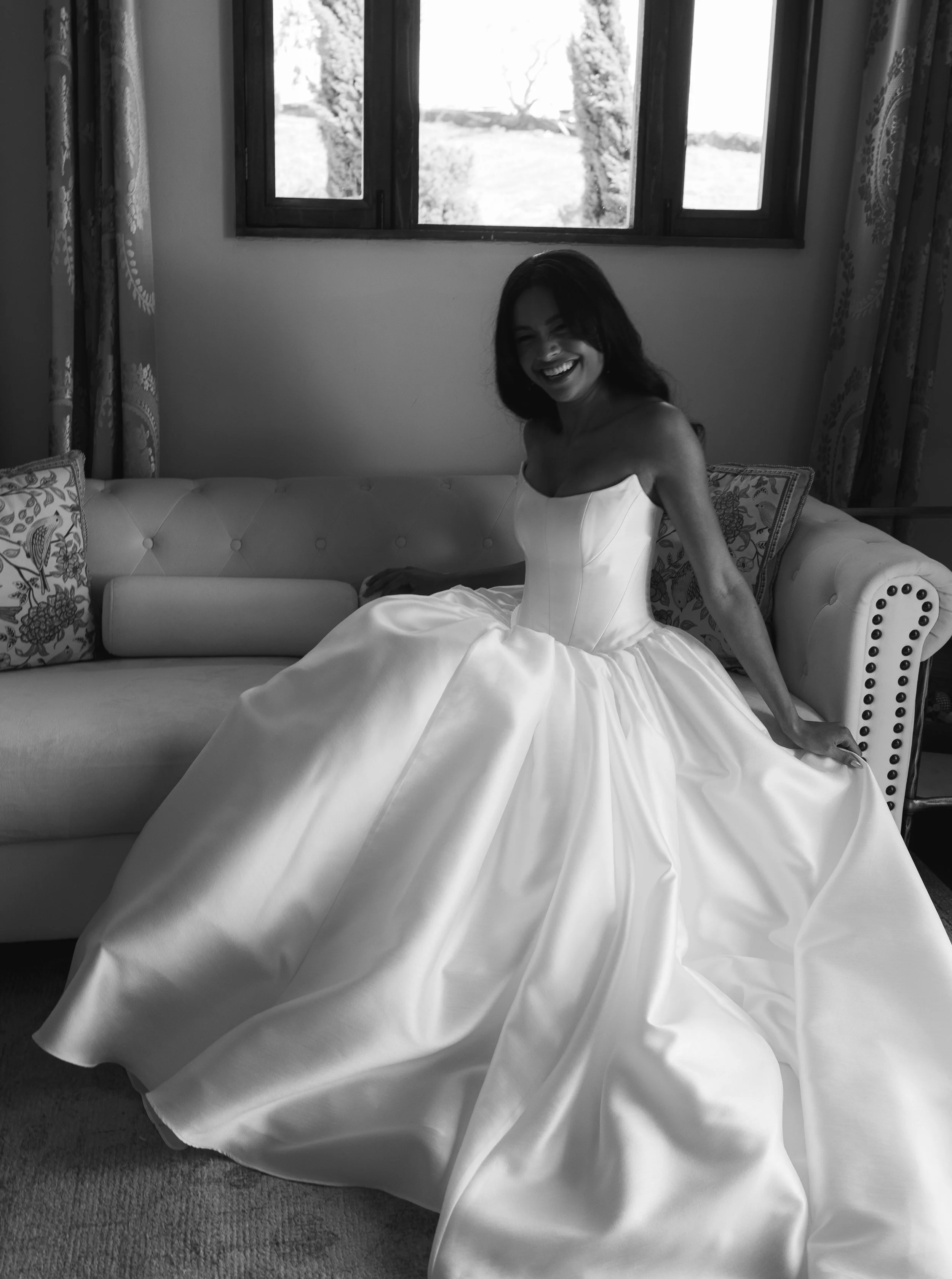 A woman in a wedding dress, sitting on a sofa near a window, smiling in a black and white photo.