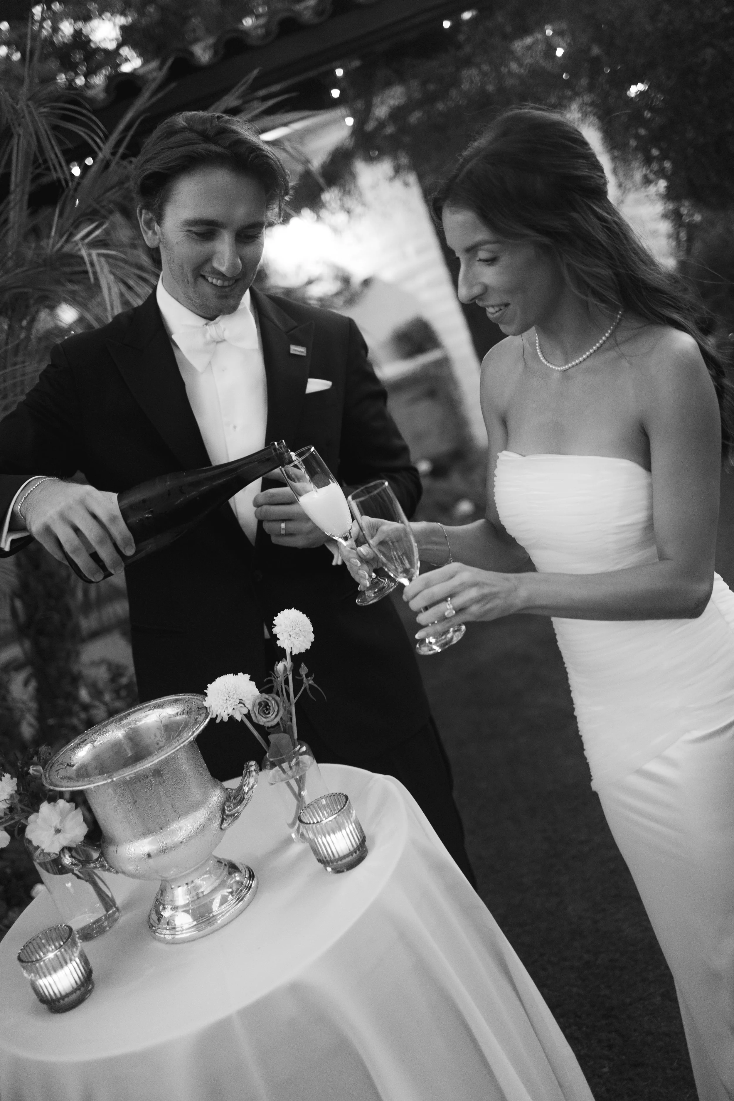A black and white photo of a wedding celebration where a groom in a tuxedo is pouring champagne into a bride's glass. The bride, in a strapless wedding dress, is holding her glass and smiling. There is a table with flowers, candles, and a champagne b