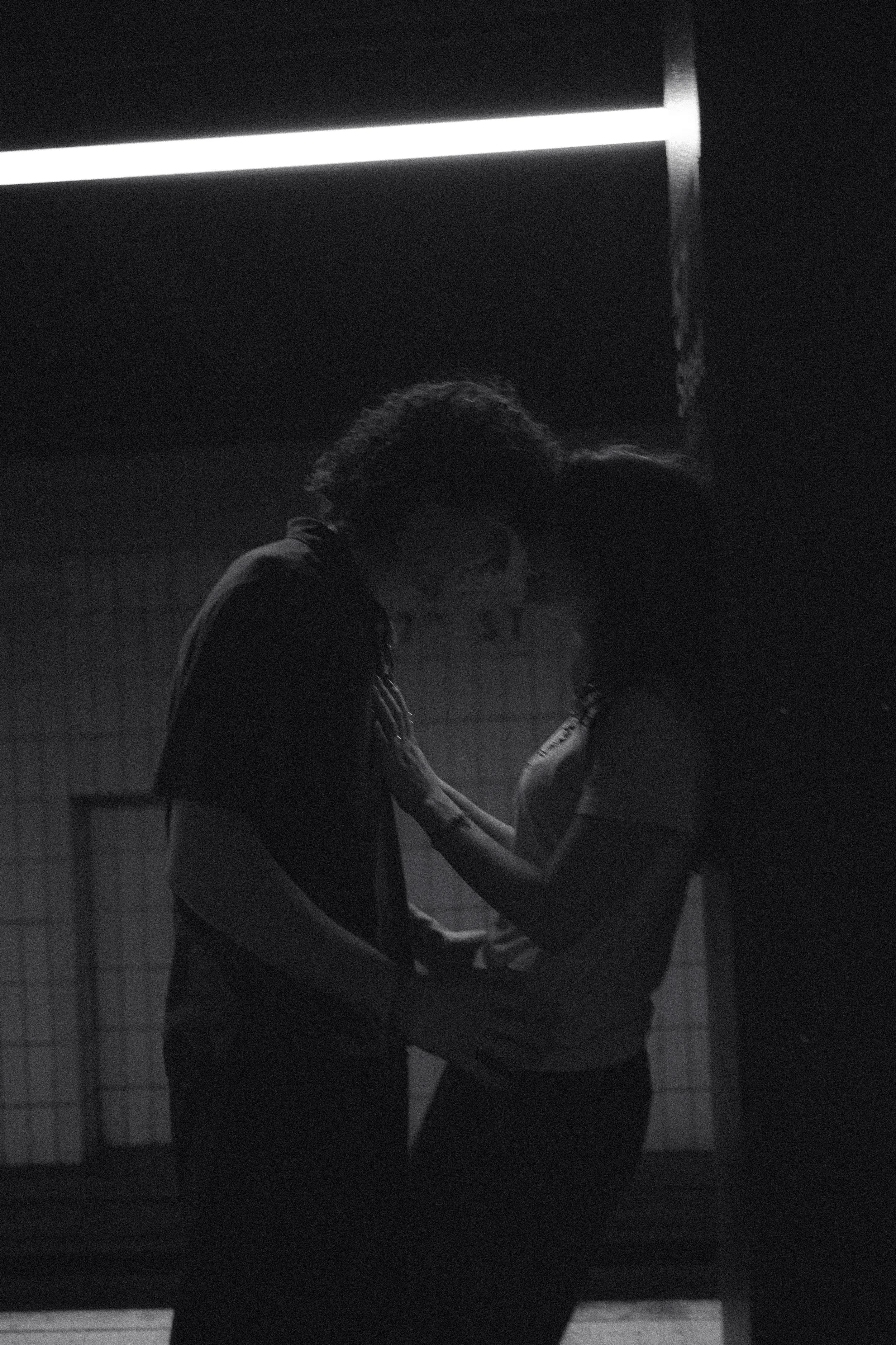 Silhouette of a man and woman in an intimate moment, holding hands and leaning towards each other in a dimly lit room.