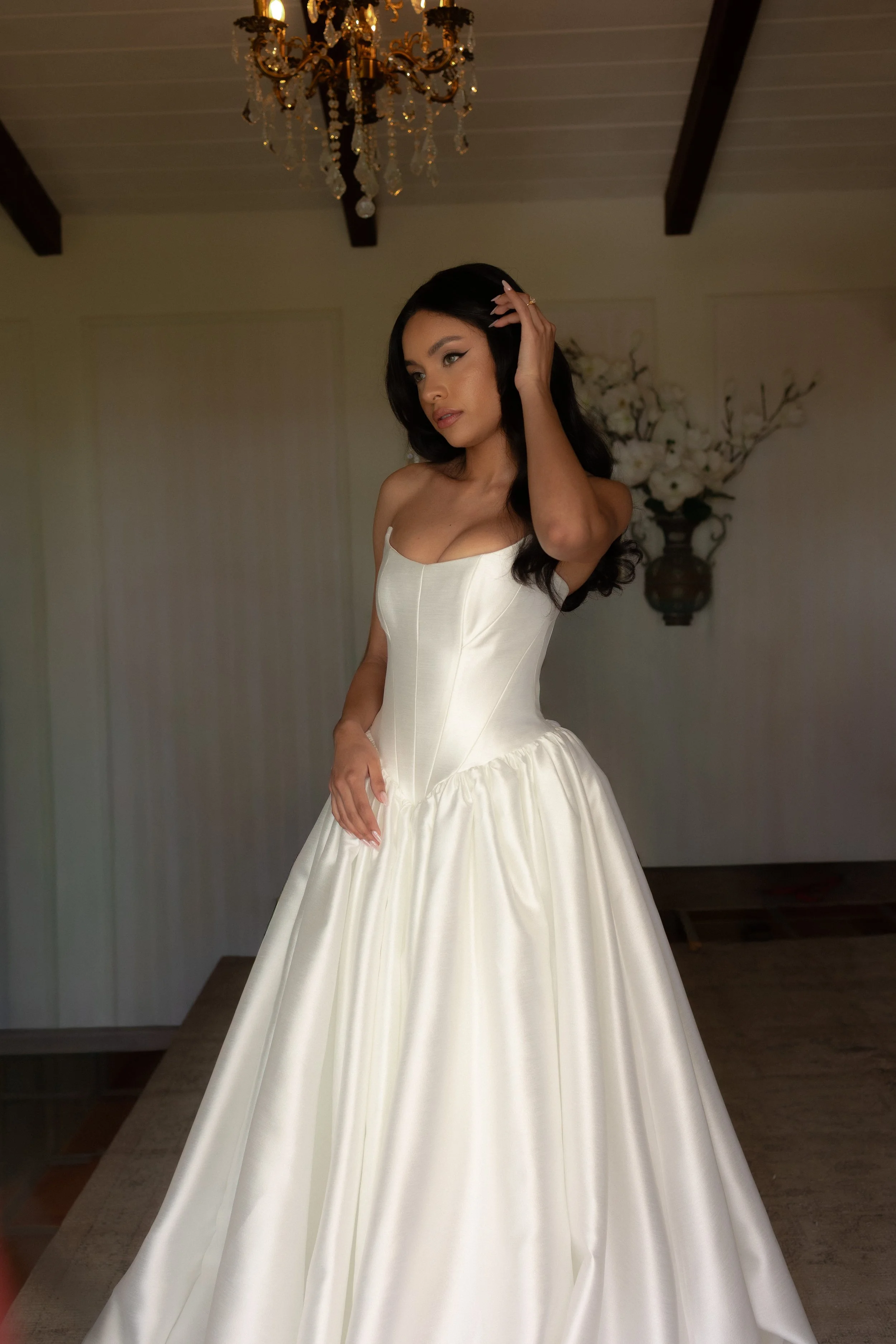 A woman wearing a strapless white wedding gown with a full skirt, standing in a room with a chandelier and floral wall decor.