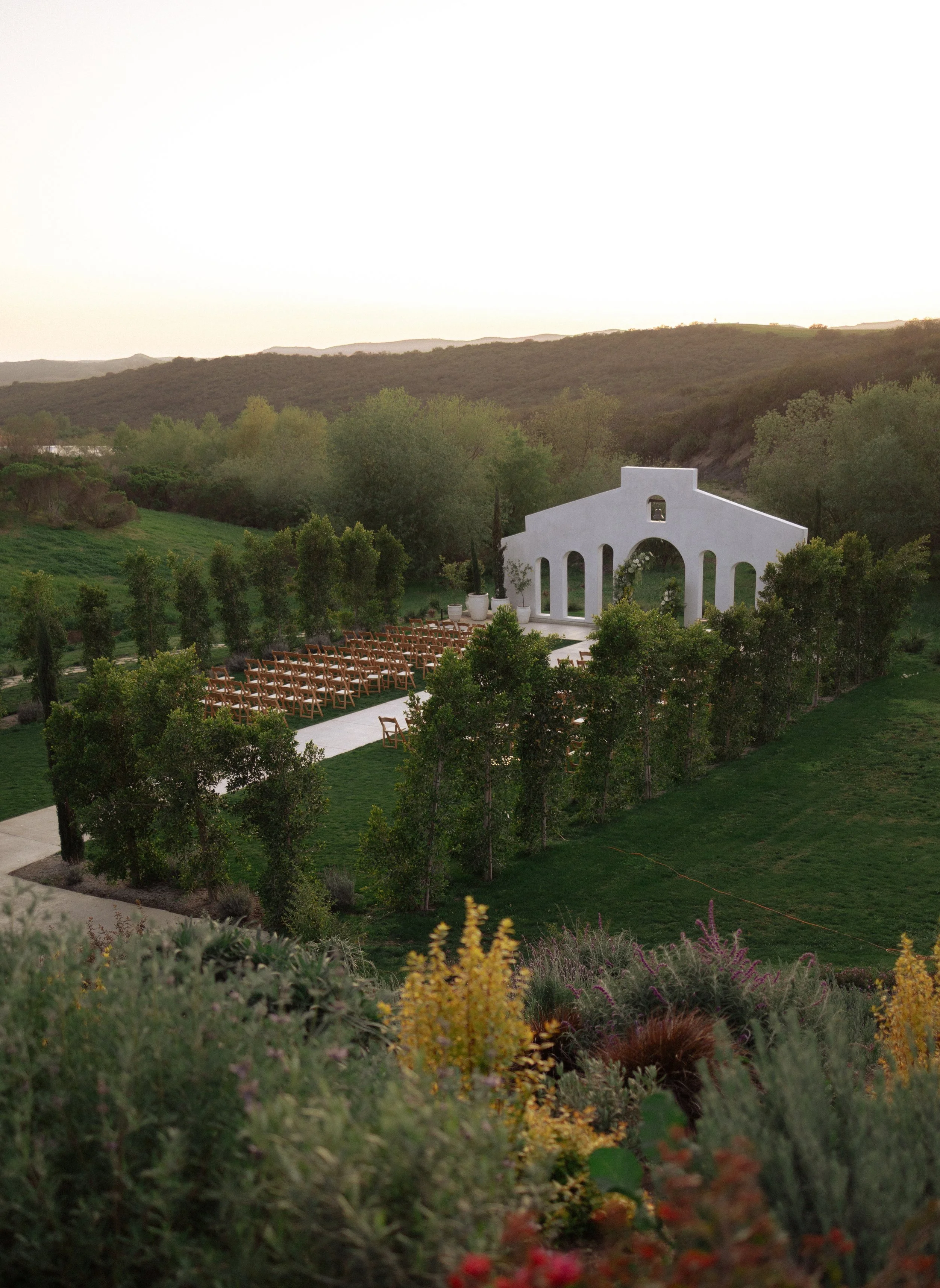 Outdoor wedding ceremony setup with wooden chairs arranged in rows facing a white chapel in a green landscape, with hills and trees in the background.