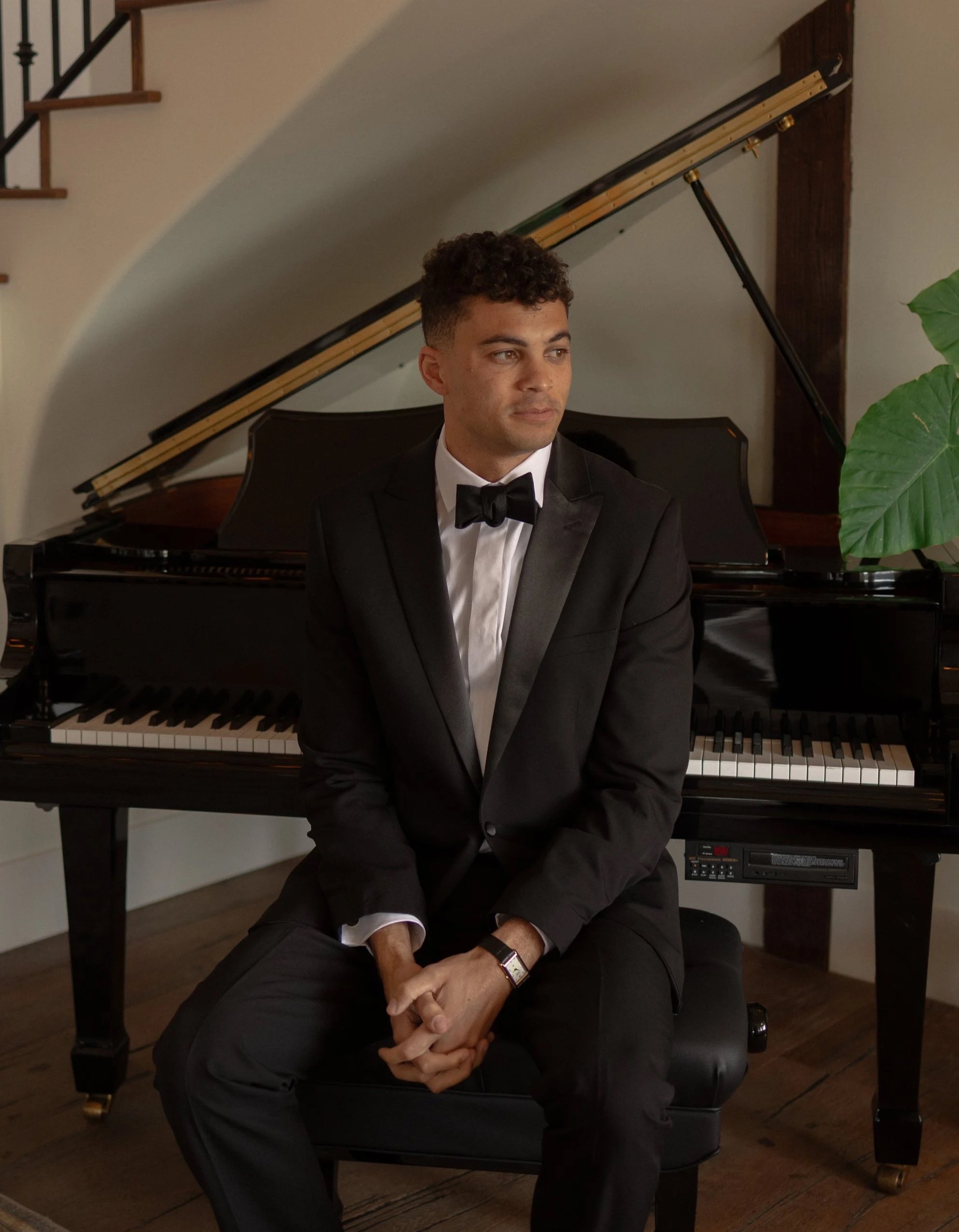 A young man in a black tuxedo with a bow tie sitting on a piano bench in front of a grand piano, inside a room with hardwood floors, a large leafy plant, and a staircase with black railings.