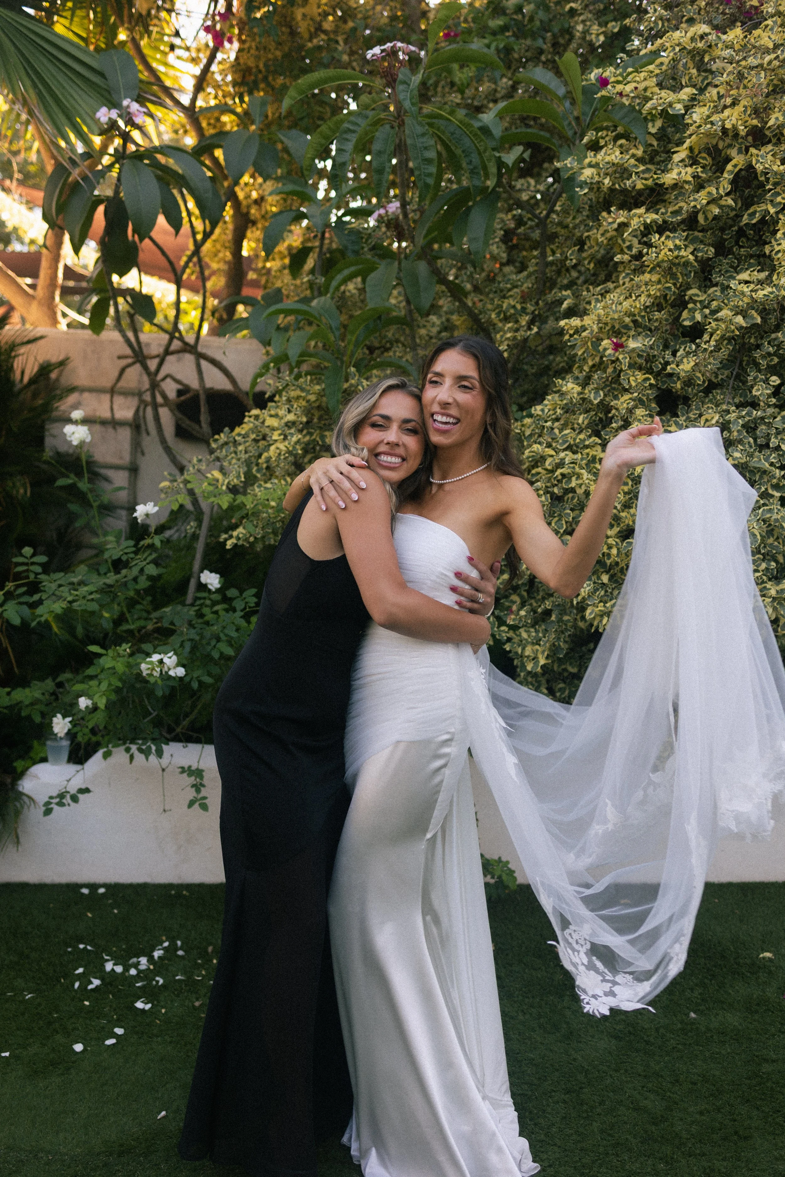 Two women hugging, one in a wedding dress and the other in a black dress, smiling outdoors in a garden with green plants and trees.