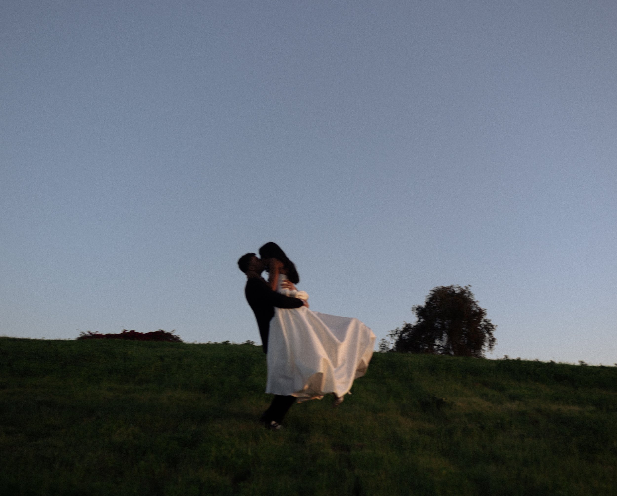 A couple dancing outdoors on a grassy hill at dusk, with the man lifting the woman who is wearing a white dress, as they share a kiss against a clear sky and a distant tree.