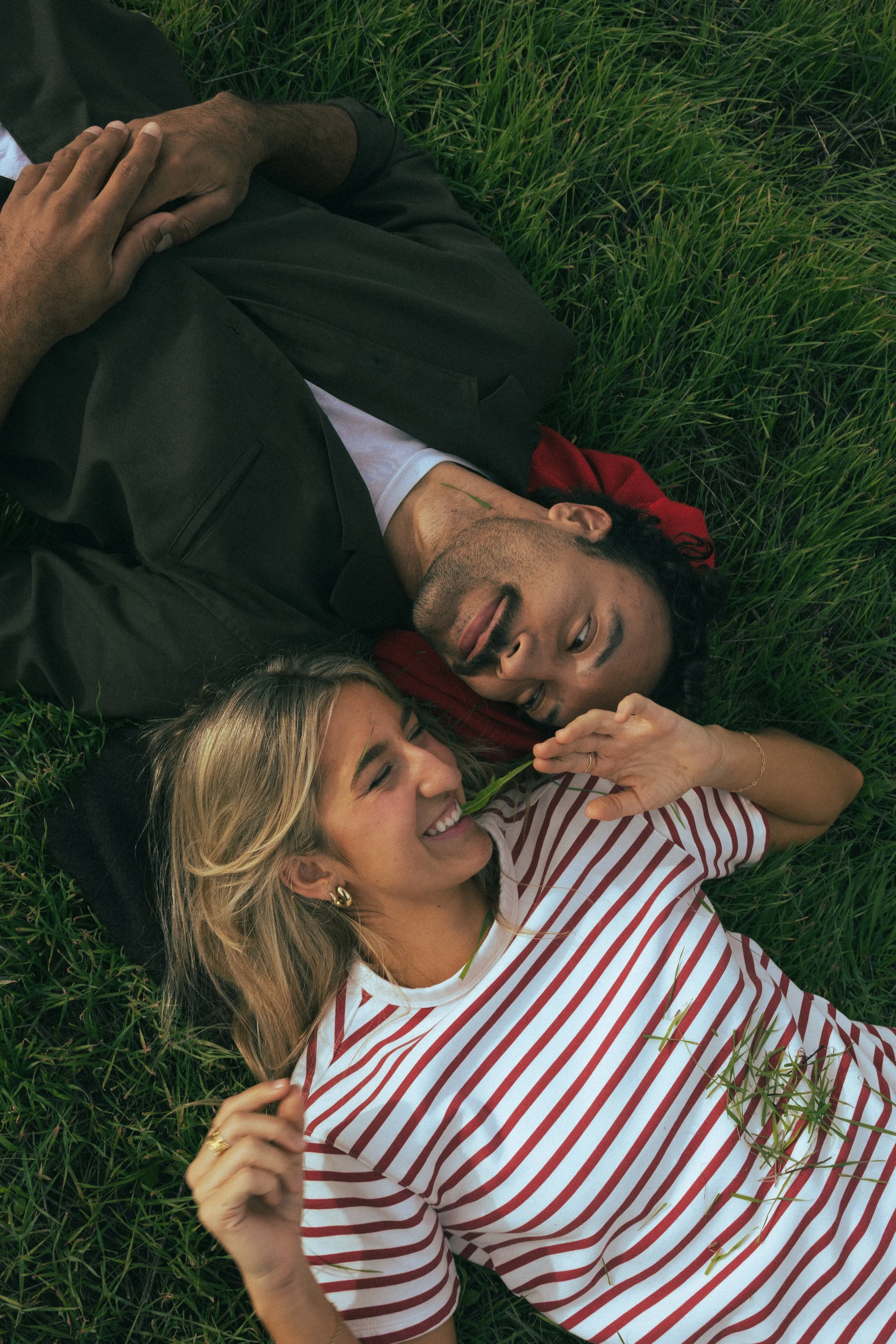 A man and woman lying on grass, smiling and playfully touching each other's faces, enjoying a moment outdoors.