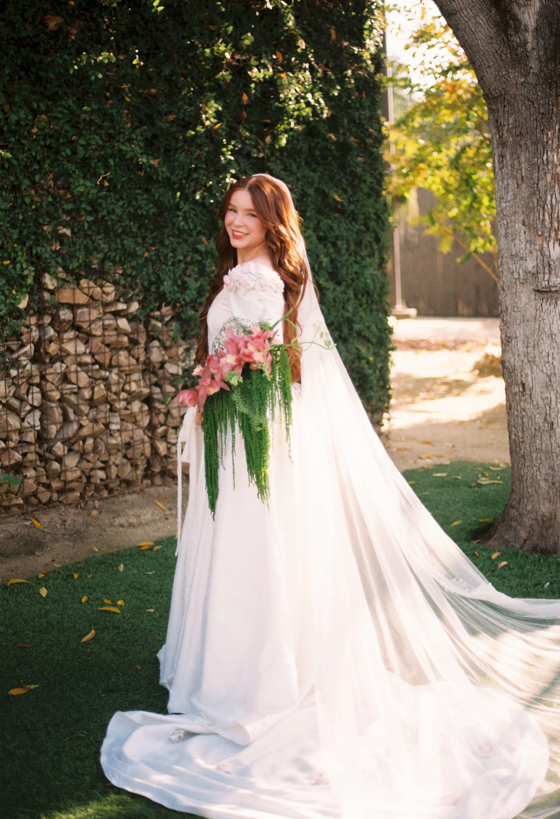 A woman in a white wedding dress holding a bouquet of pink flowers and greenery, standing outdoors near a tree and a hedge, with sunlight filtering through the leaves.
