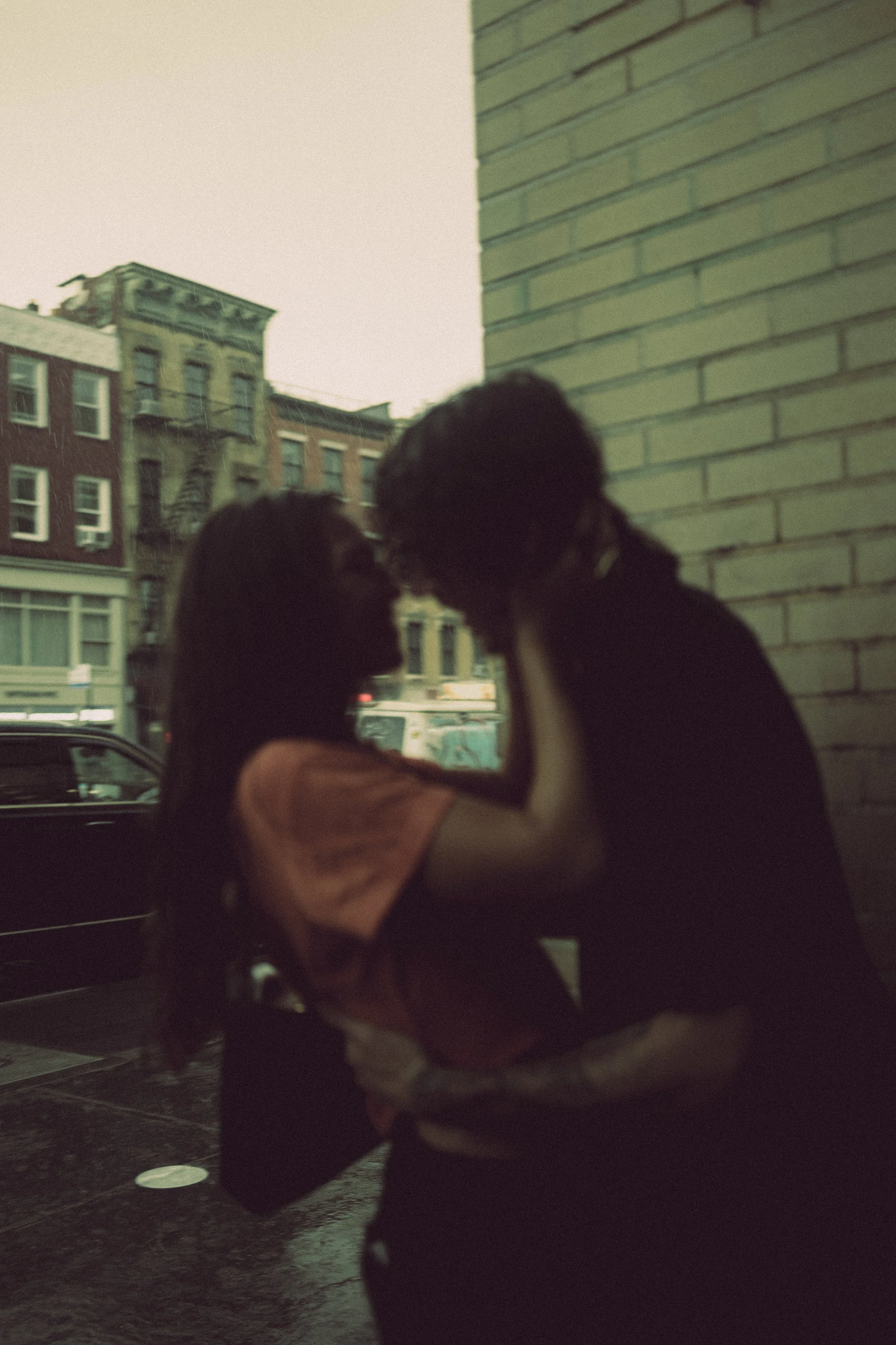 A couple stands close together outdoors in front of a brick wall, about to kiss, with city buildings and parked cars in the background.