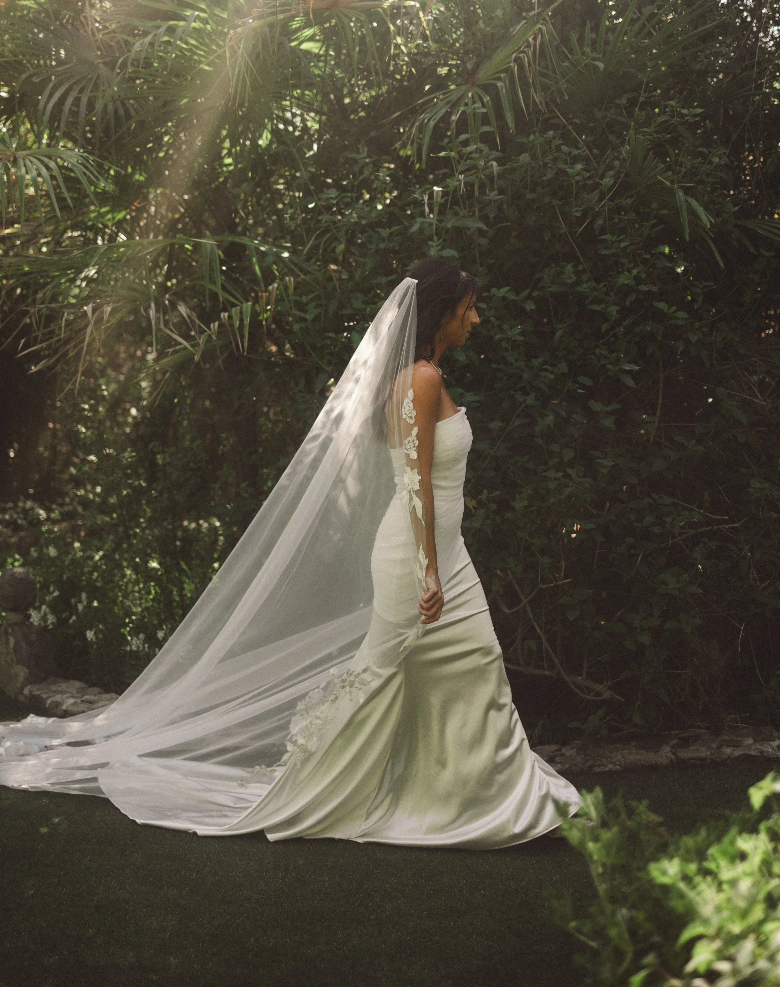 A woman in a white wedding gown with a long train and lace details, wearing a veil, standing outdoors amidst lush green foliage.