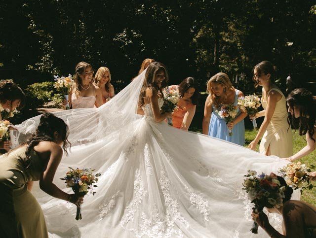Bride in a white wedding gown and veil surrounded by bridesmaids holding bouquets, outdoors with trees in the background.
