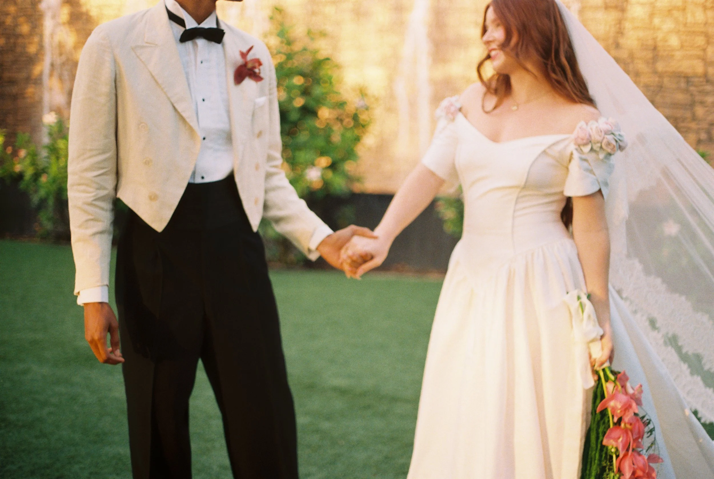 A bride and groom holding hands outdoors during their wedding ceremony, with the bride holding a bouquet of flowers and smiling.