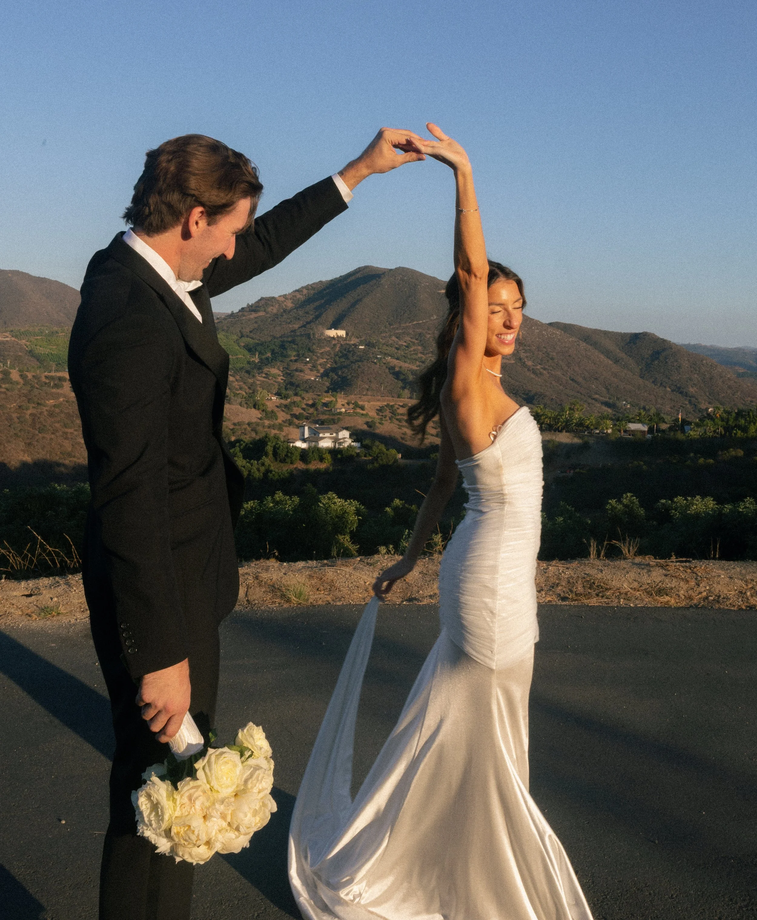 A man in a black suit with a bouquet of white roses dances with a woman in a white wedding dress outdoors on a sunny day with mountains in the background.