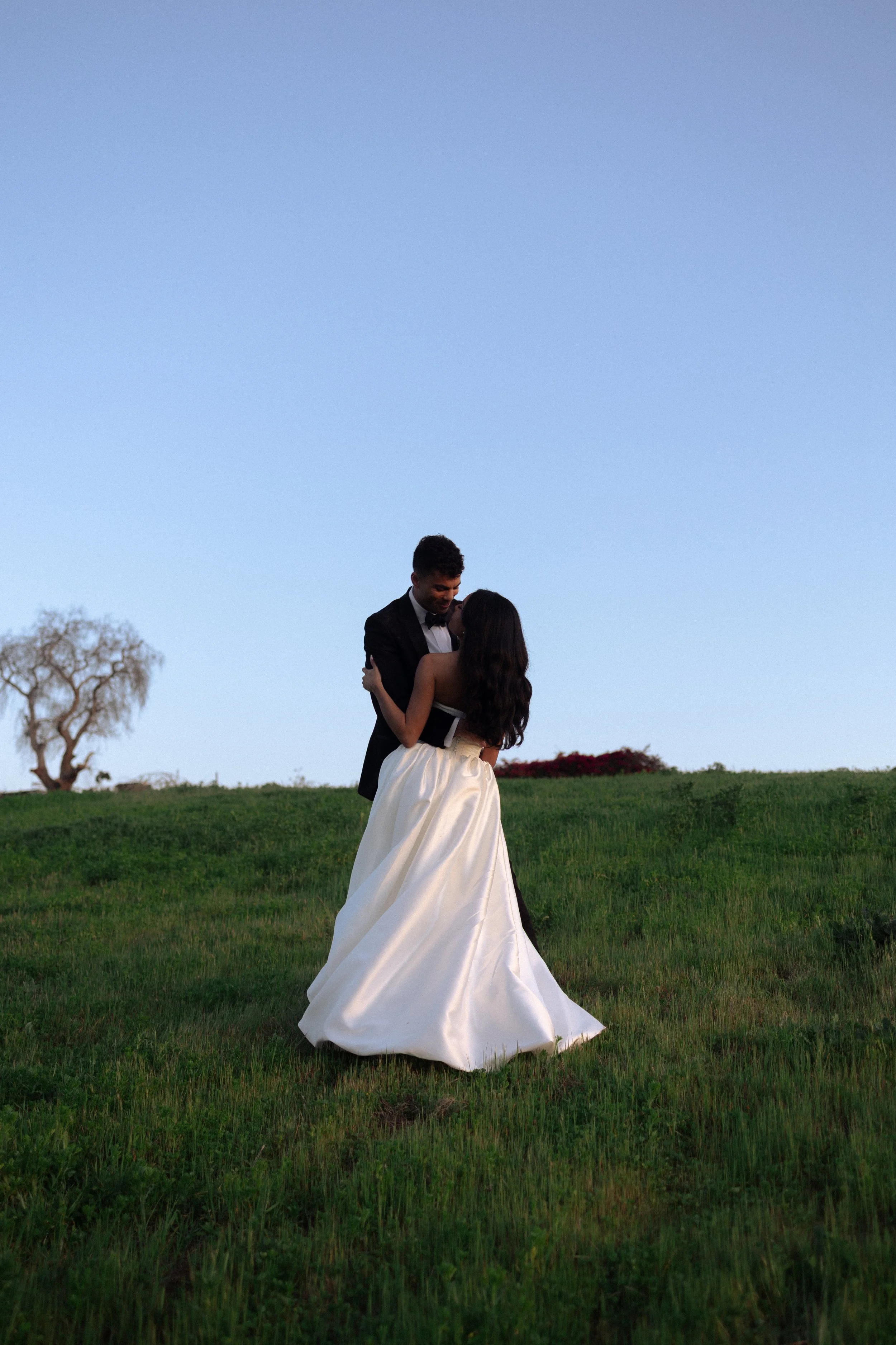 A couple dancing outdoors on a grassy hill, with a clear blue sky in the background. The woman is wearing a white wedding gown and the man is dressed in a black tuxedo.