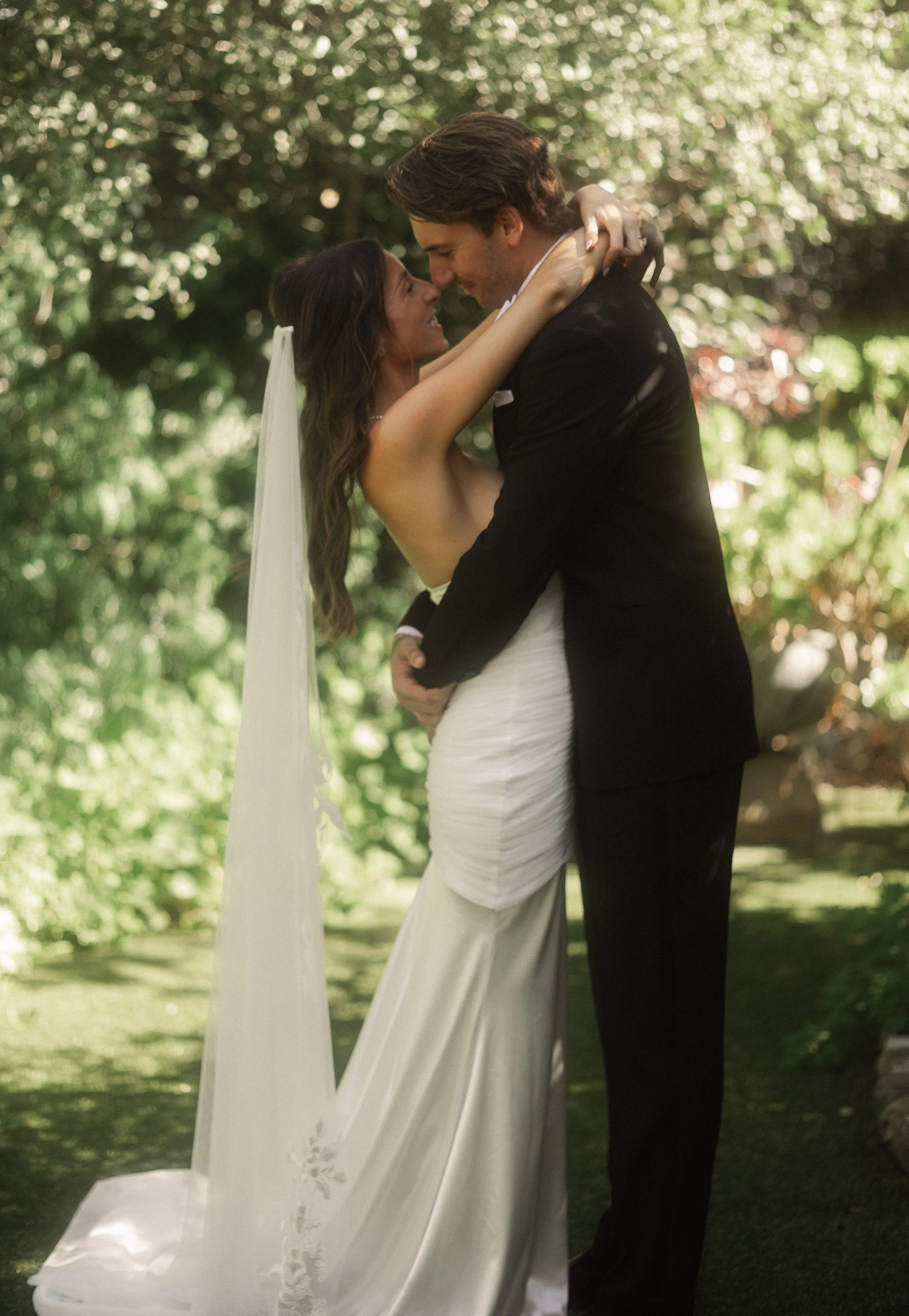 A bride and groom embracing outdoors on their wedding day, surrounded by greenery.