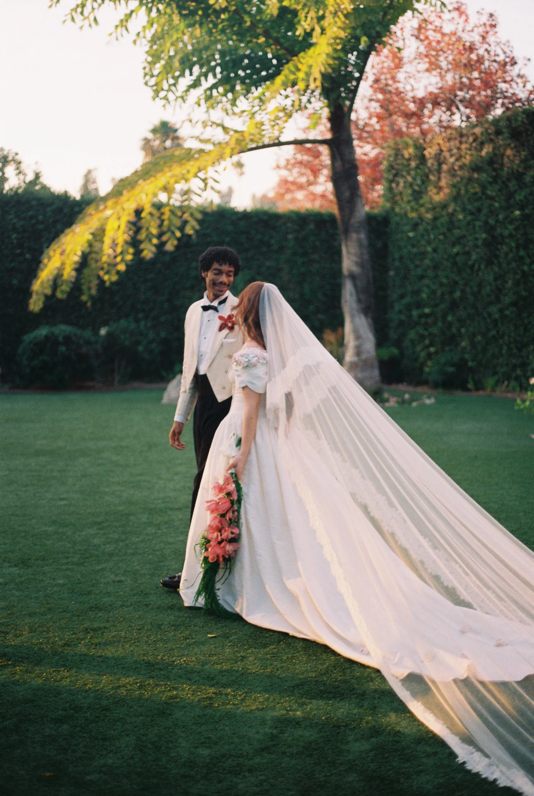 A bride and groom on grass in a garden, with the bride holding a pink floral bouquet and wearing a lace wedding dress and long veil, while the groom is in a tuxedo with a bow tie, smiling at each other.