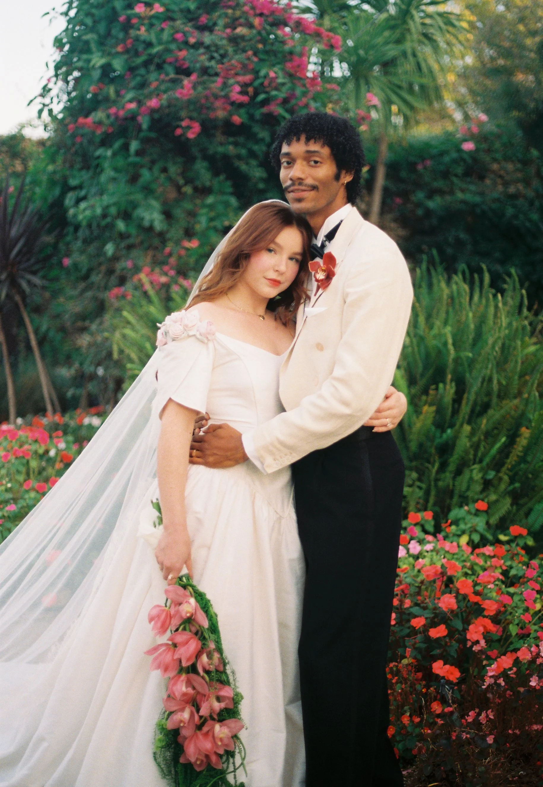 A bride and groom standing closely together outdoors in a lush garden with pink and red flowers, the bride holding a cascading bouquet of pink flowers, and the groom wearing a cream tuxedo jacket with a red orchid boutonniere.
