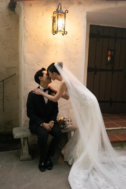Bride leaning over to kiss groom on the nose, both dressed in wedding attire, sitting on a gray stone bench outside near an illuminated lamp and a black gate.