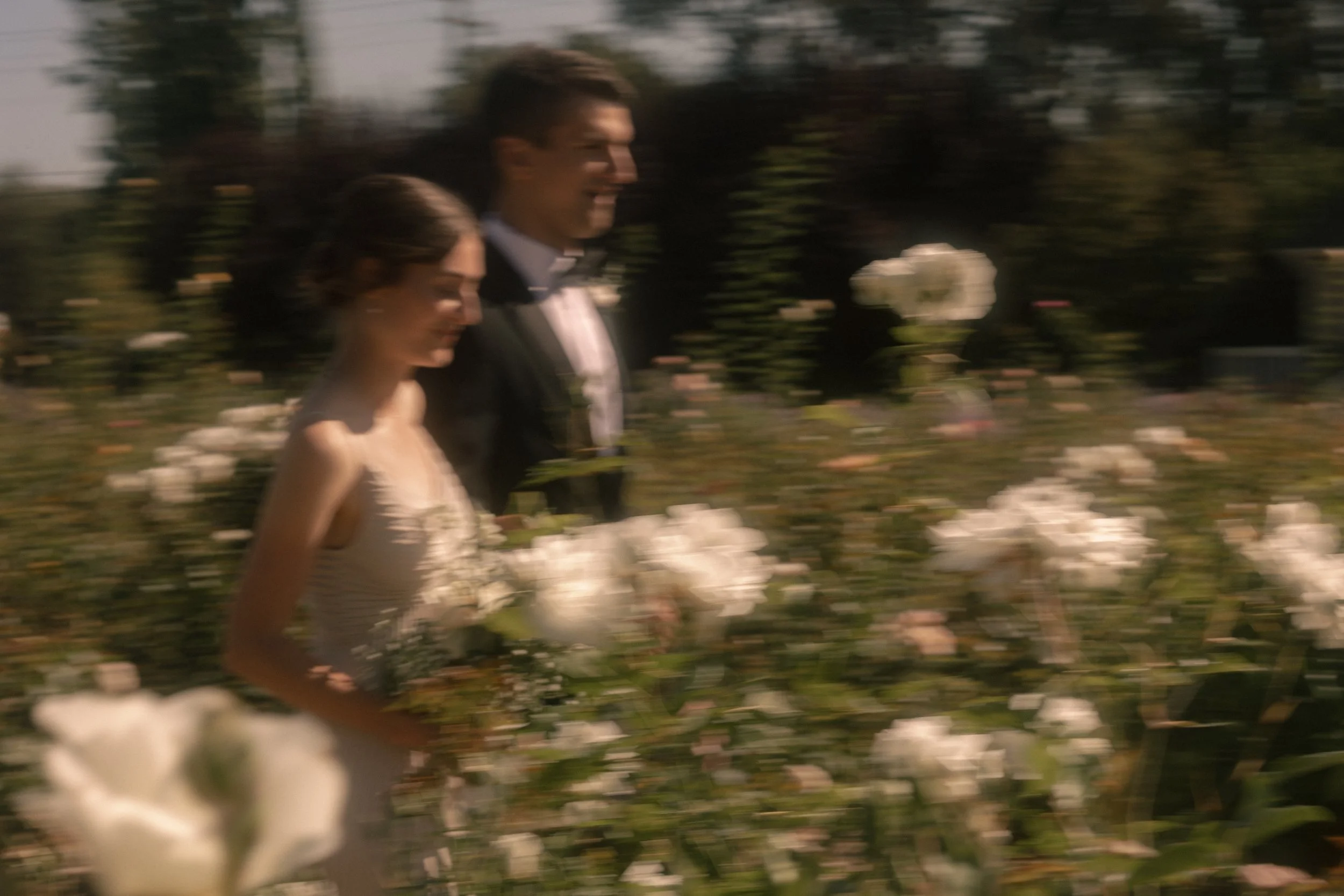 Blurred photo of a bride and groom walking through a garden with white flowers.