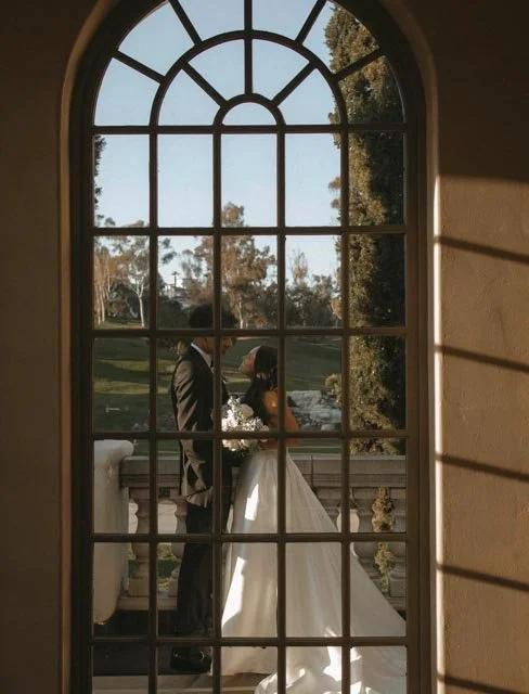 A couple dressed in wedding attire standing together outside on a balcony, seen through a large arched window with grid panes, in a park-like setting.