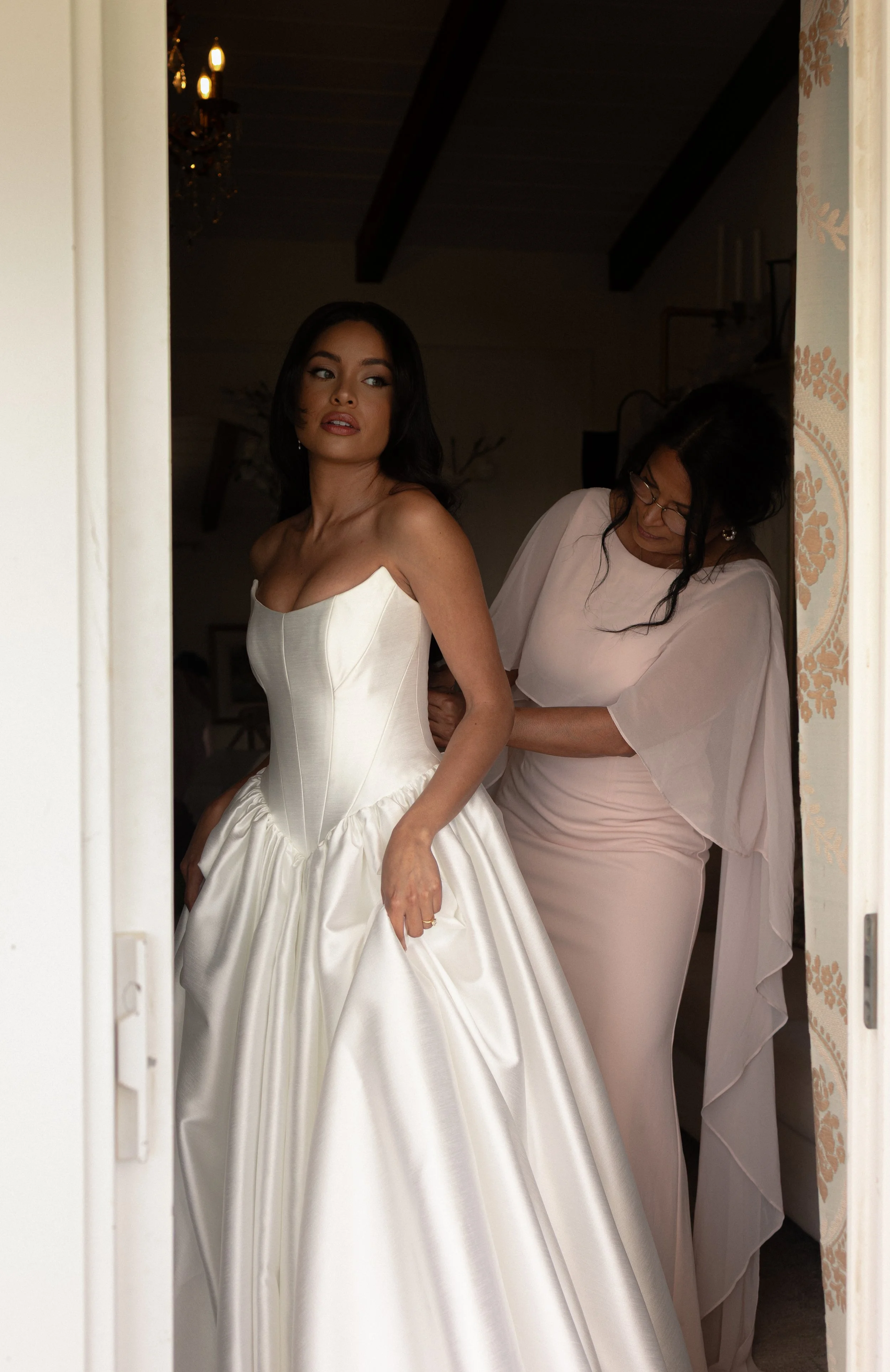 A woman in a wedding dress being assisted by another woman in a light pink dress.