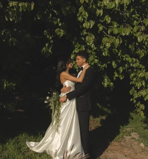 A bride and groom sharing a kiss outdoors under a tree at night, with the bride holding a bouquet of flowers.