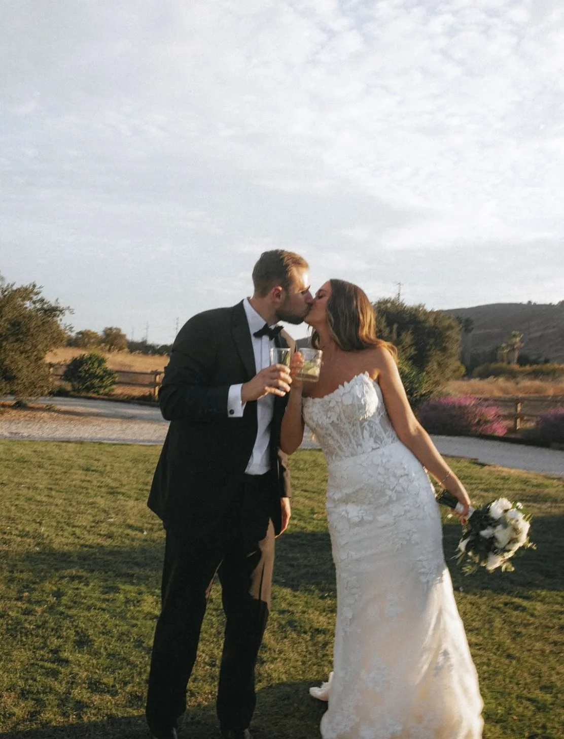 A couple in wedding attire kissing outdoors, holding drinks, with the bride holding a bouquet, on a grassy area near a fence and trees under a partly cloudy sky.