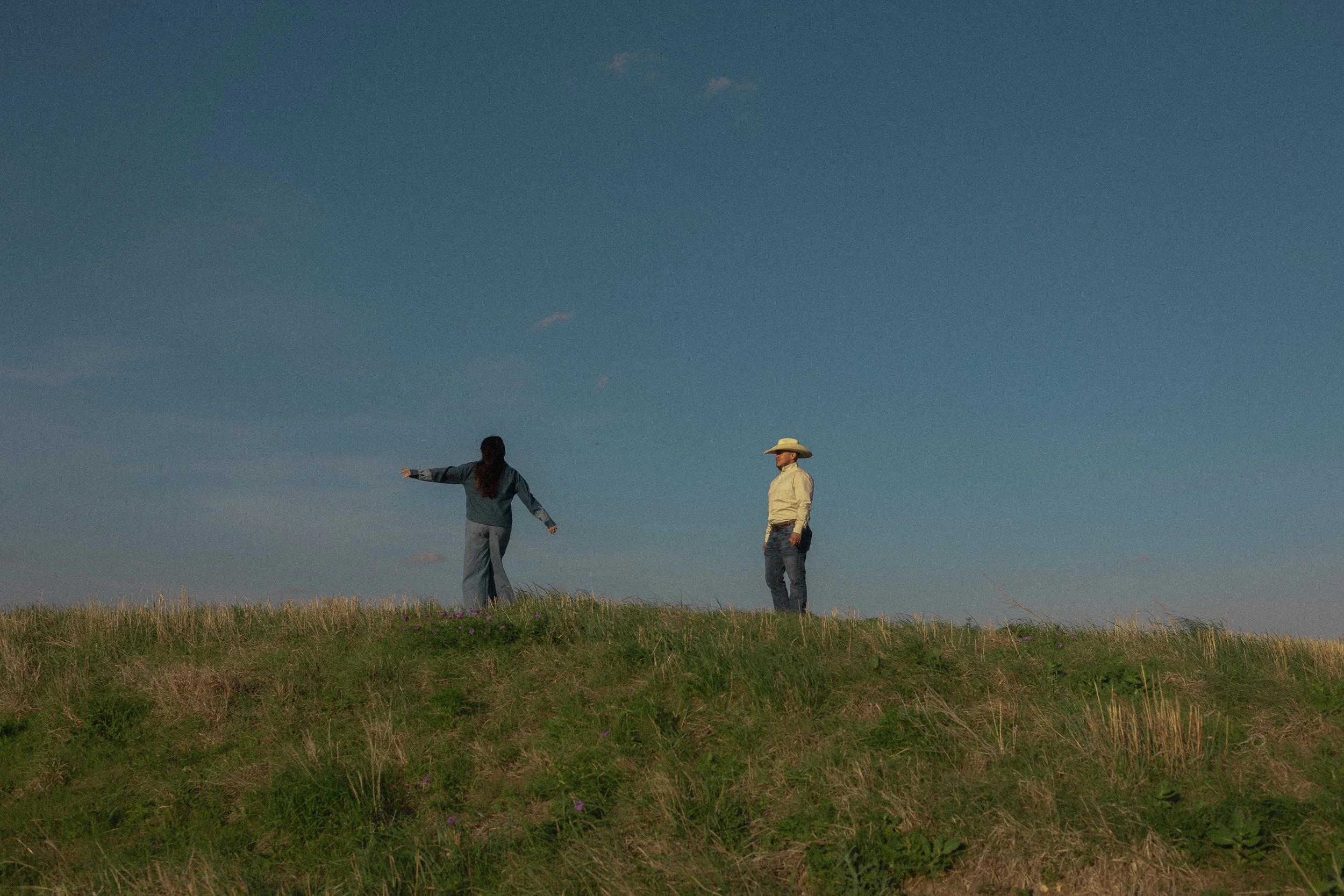 Two people standing on a grassy hill under a clear blue sky, one woman with long hair wearing jeans and a jacket with arms outstretched, and a man wearing a cowboy hat and jeans standing nearby.
