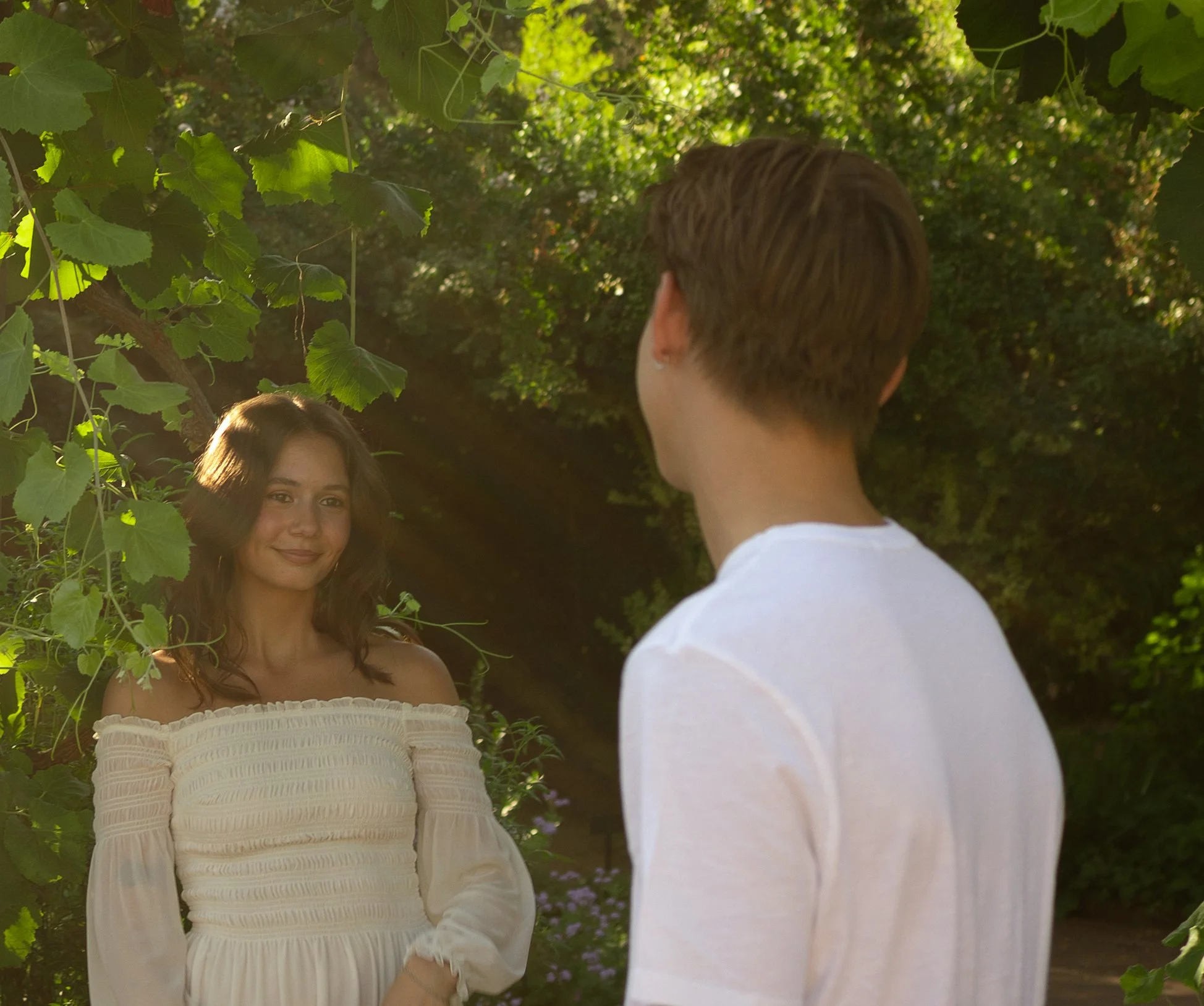 A woman with wavy brown hair and a white off-shoulder dress standing outdoors among green foliage, smiling, while a man with short brown hair in a white shirt faces her with his back to the camera, in a natural setting with sunlight.
