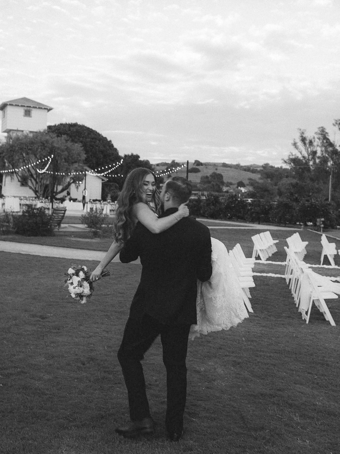A joyful woman in a dress being lifted by a man in a suit at an outdoor wedding reception, with chairs and string lights in the background.