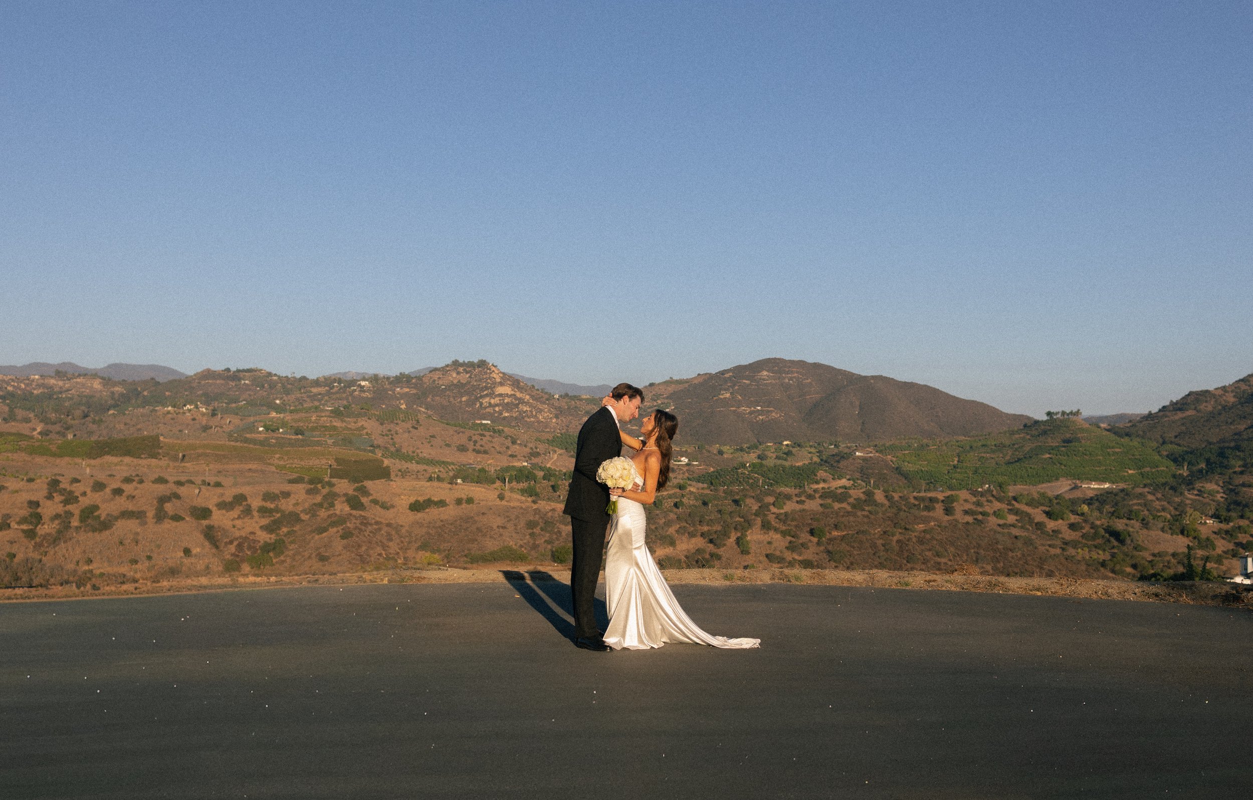 A bride and groom standing on a flat surface outdoors, embracing with mountains and a clear sky in the background.
