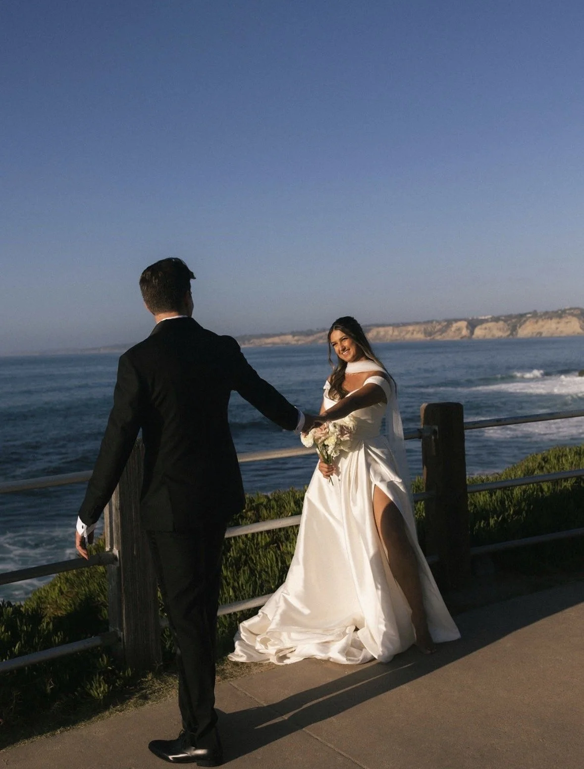 A bride and groom holding hands by the ocean at sunset, with cliffs in the background.