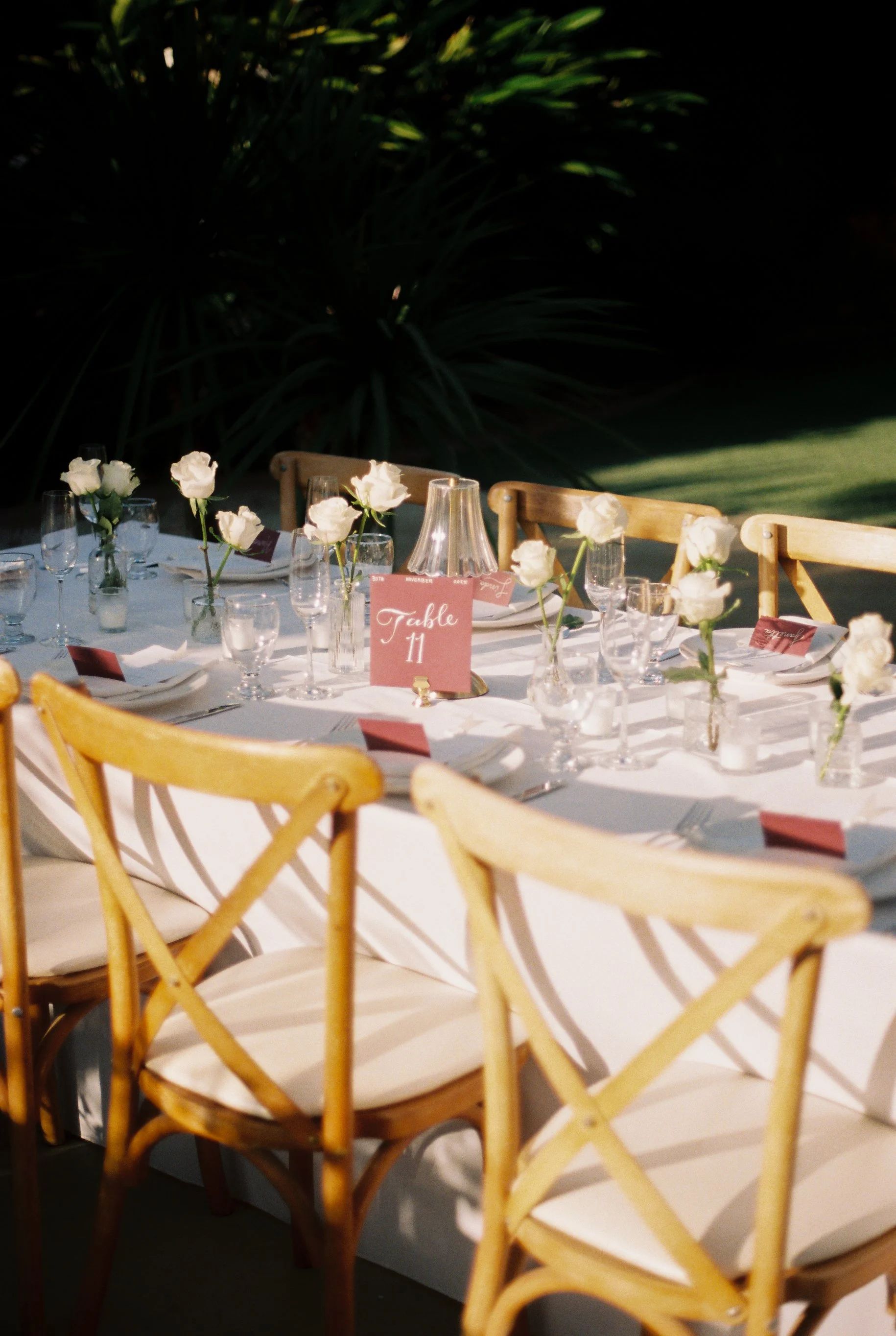 Outdoor wedding reception table decorated with white roses in small glass vases, pink place cards, water glasses, white napkins, and a pink table sign labeled 'Table 11'. Wooden chairs surround the table, with sunlit greenery in the background.