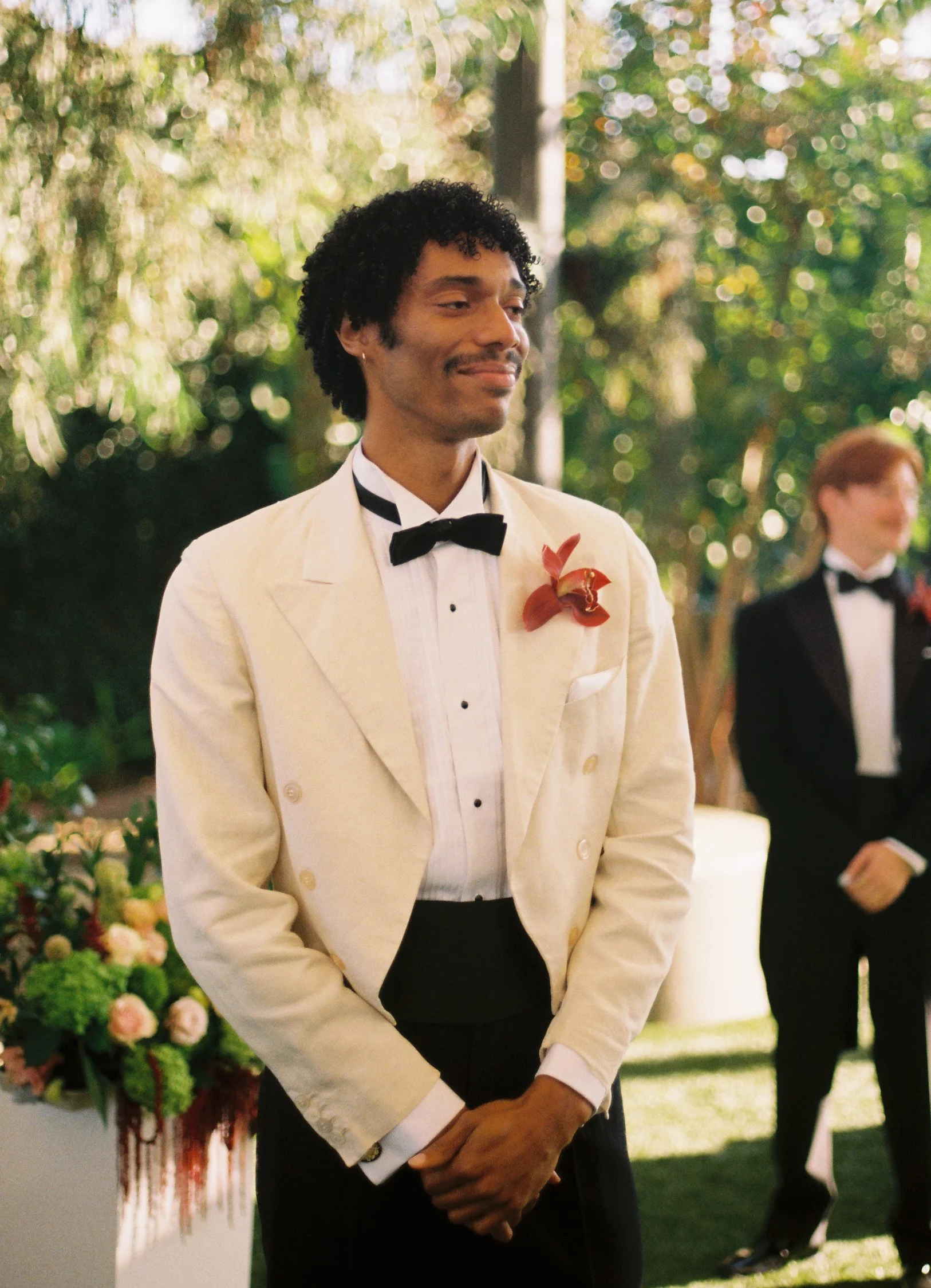 A man in a white tuxedo with a black bow tie and a red flower boutonniere, standing attentively at an outdoor wedding ceremony.