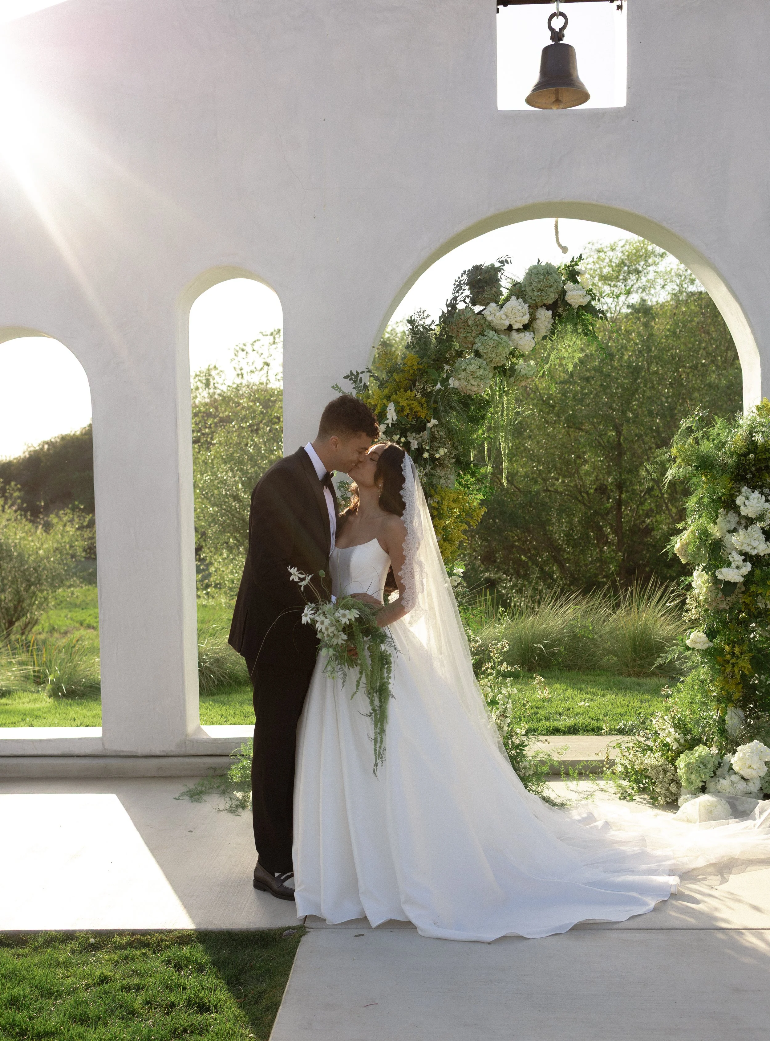 A bride and groom kissing under a floral arch outside during a wedding ceremony.