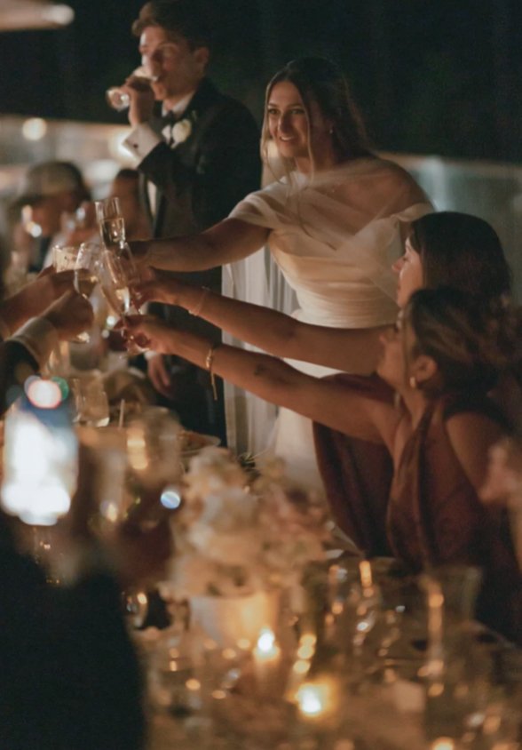 A woman in a white dress is smiling and reaching across a table to toast with another woman at a formal event or wedding reception, with a man in a tuxedo drinking a beverage in the background.
