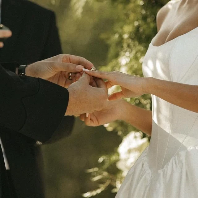 A couple exchanges wedding vows, holding hands, with a person in a black suit and a bride in a white dress outdoors.