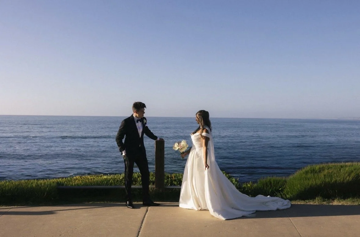 A bride and groom in wedding attire standing by the ocean, facing each other, with the bride holding a bouquet of flowers on a pathway beside green grass.