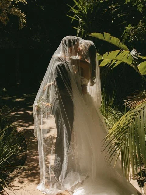 Bride and groom standing close under a wedding veil outdoors surrounded by tropical plants.