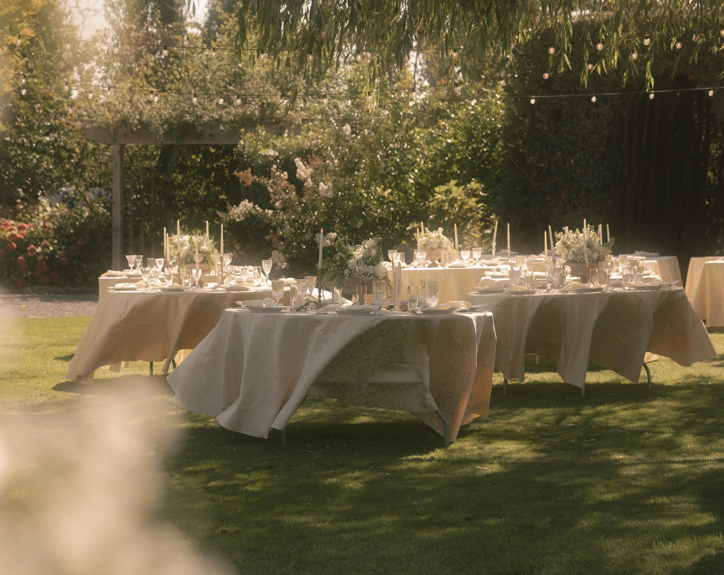 An outdoor dining setup with multiple round tables covered in white tablecloths, decorated with floral centerpieces and lit Candles, set in a garden area with green grass and trees, and string lights overhead, during daylight.