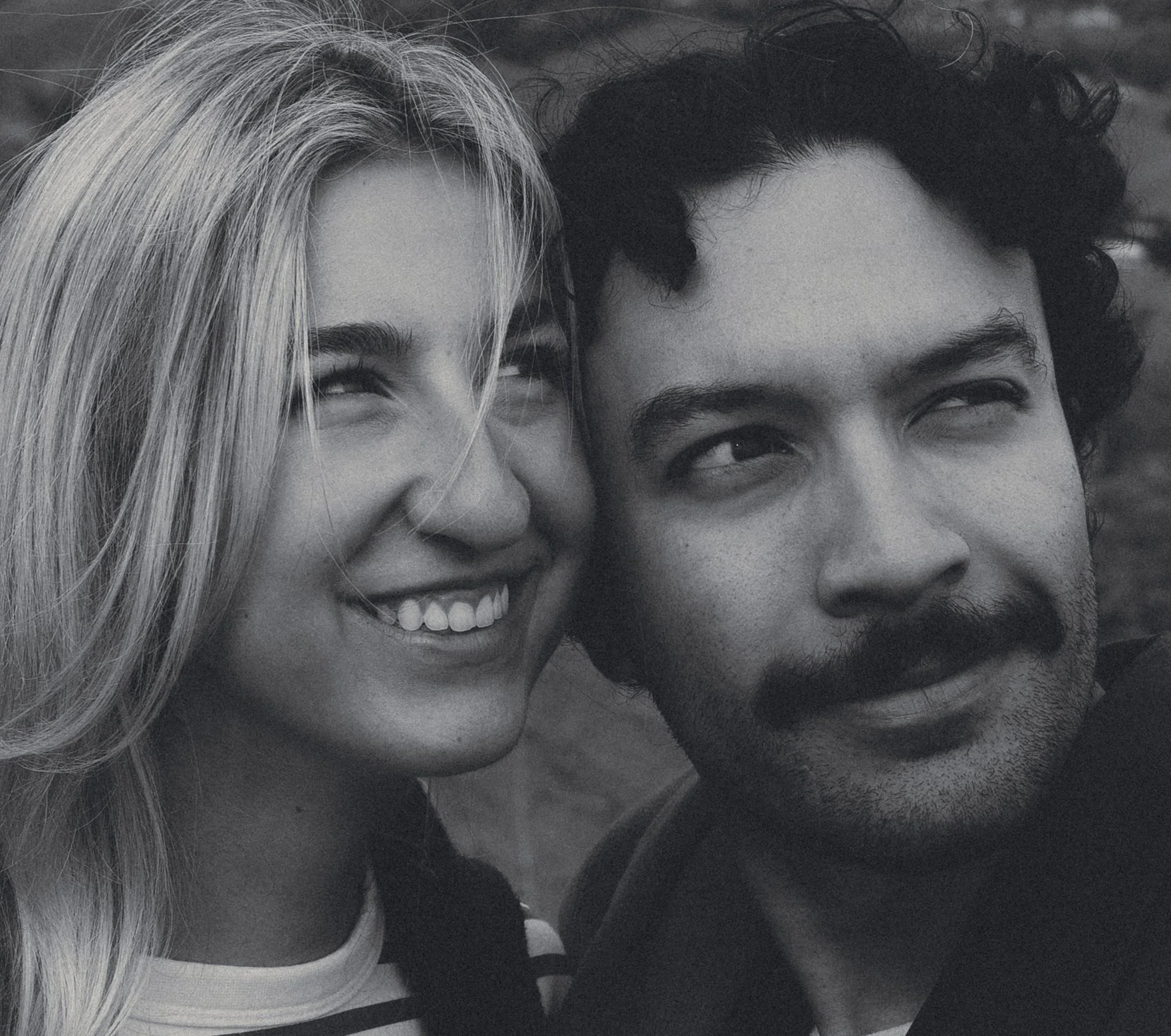 Close-up black and white photo of a smiling woman with long blonde hair and a man with curly dark hair and a mustache, both looking to the side. best wedding photographer in dallas