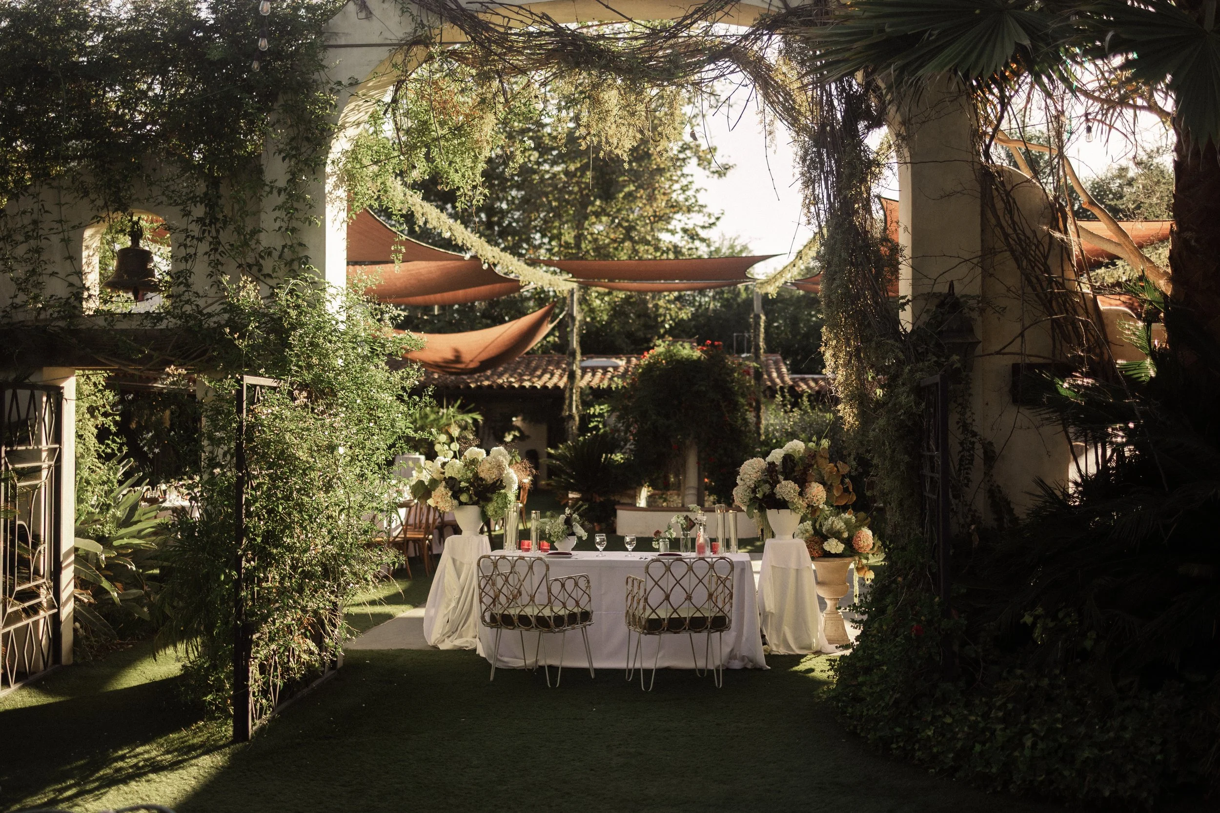 A beautifully decorated outdoor garden with a white table set for a formal event, surrounded by lush greenery, flowers, and covered with brown shade canopies.