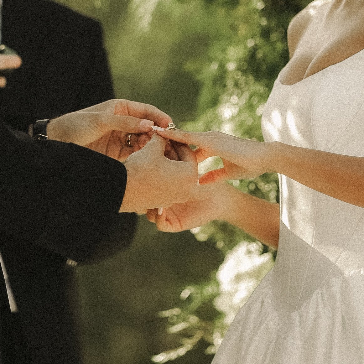 A bride and groom exchanging rings outdoors, with the groom's hands placing a ring on the bride's finger.