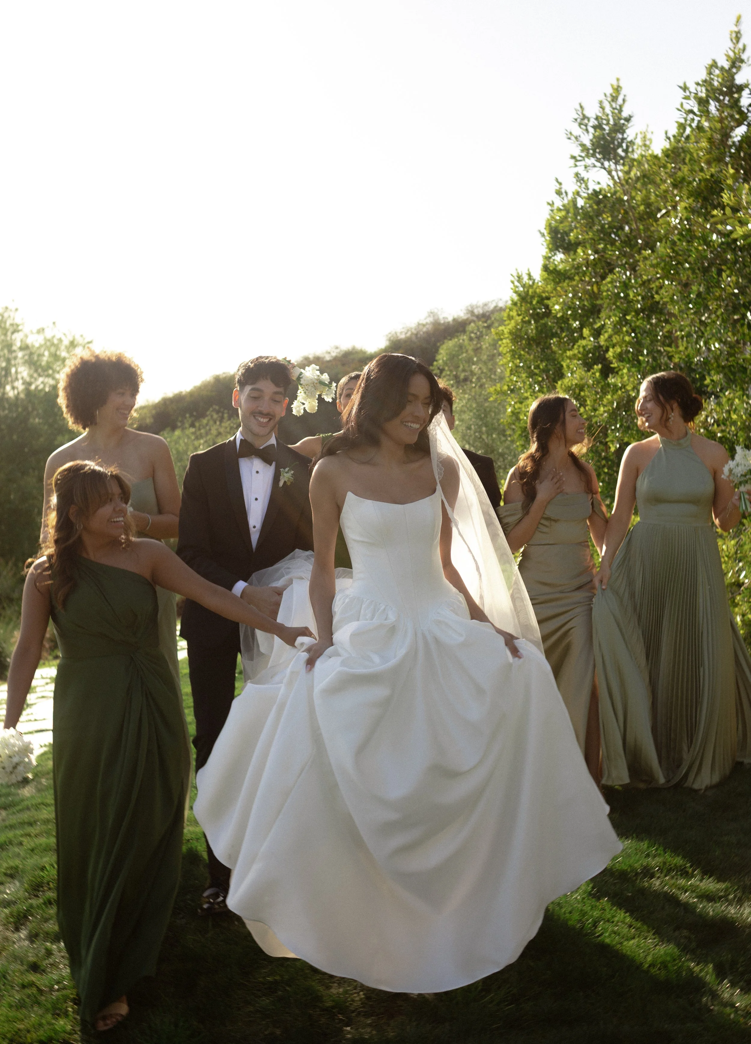 A bride in a white wedding gown walking outdoors with her wedding party, including bridesmaids in green dresses and groomsmen in tuxedos, under green trees on a sunny day.