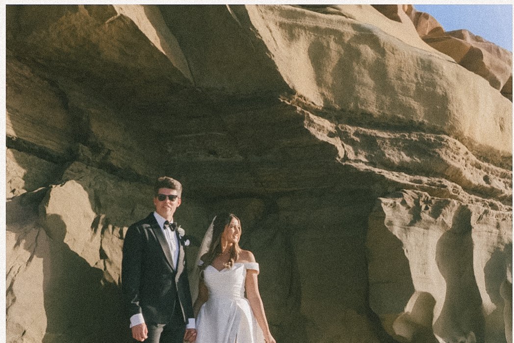 A bride in a white wedding dress and a groom in a black tuxedo holding hands, standing outdoors against large rock formations.