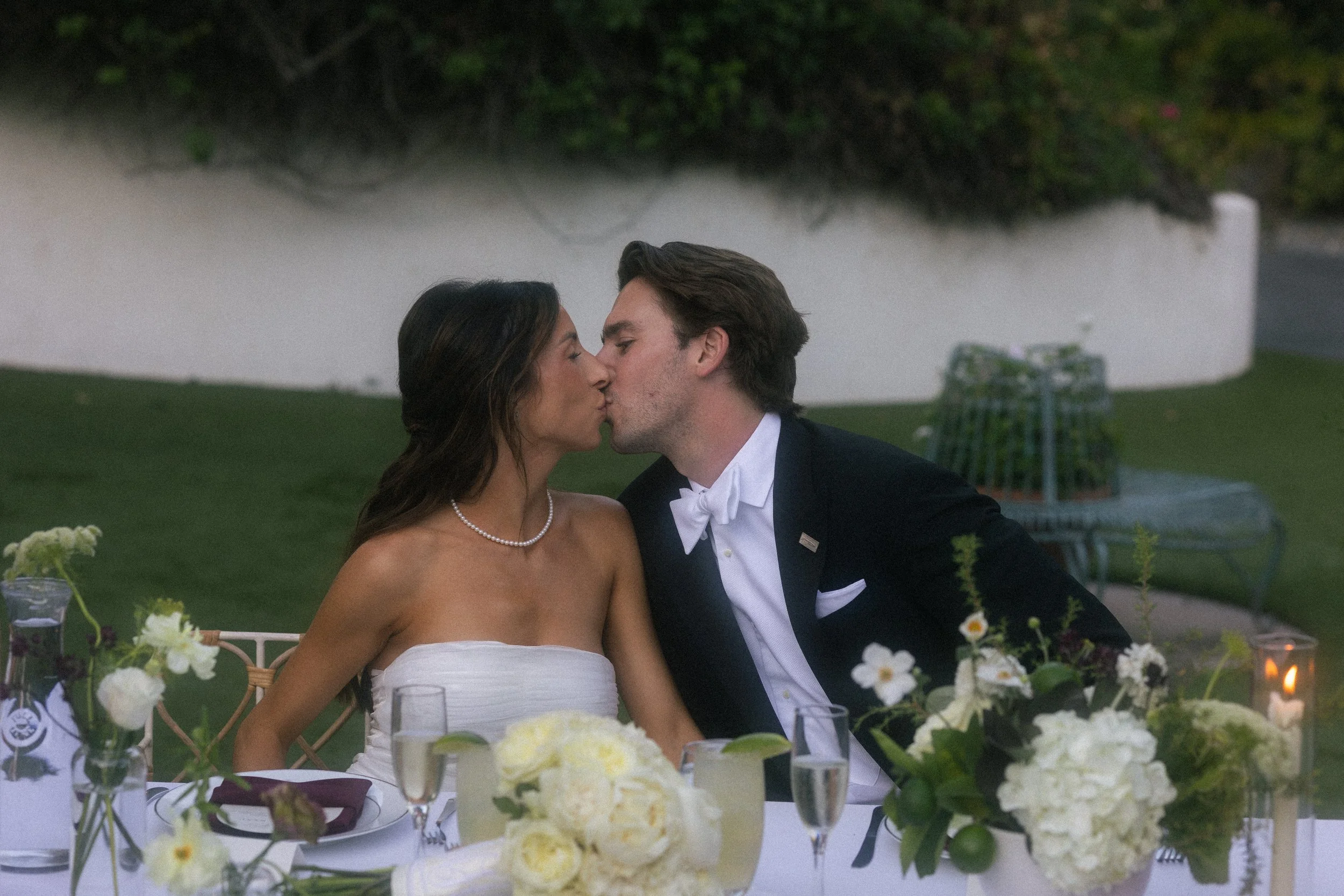 A couple in wedding attire sharing a kiss at a decorated outdoor table.