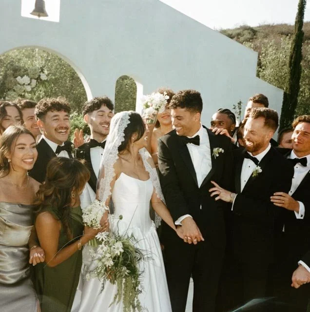 Wedding celebration outdoors with bride and groom holding hands surrounded by friends.
