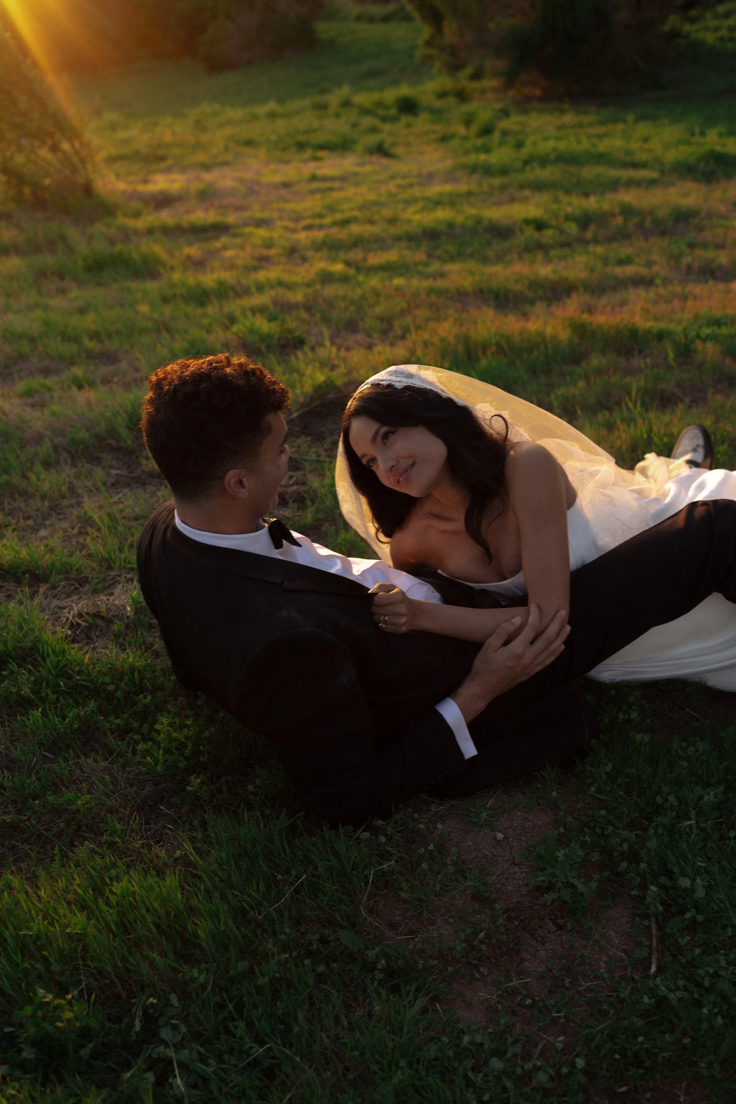 A bride and groom sitting on grass in a field during sunset, smiling and gazing at each other.