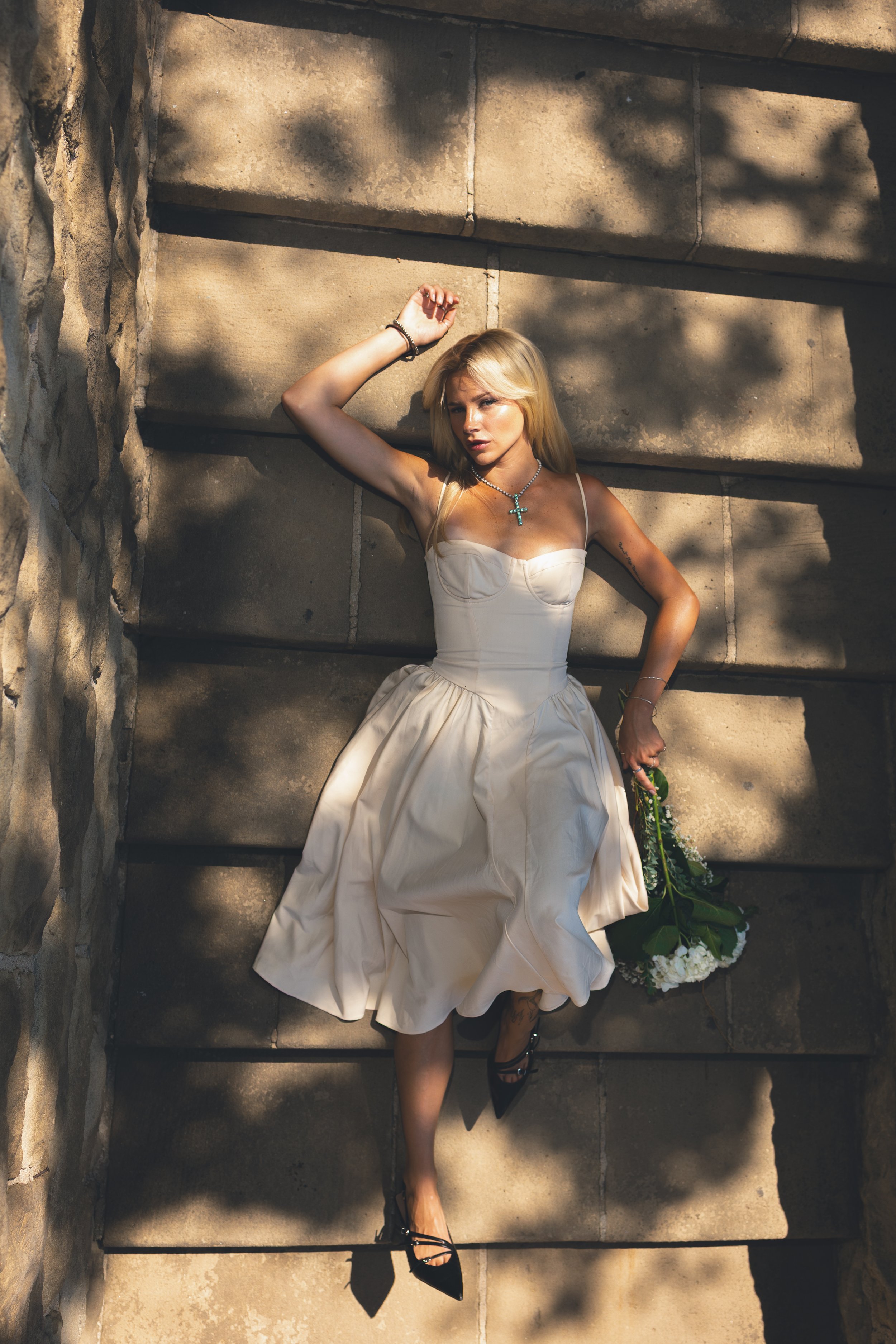 A woman in a white dress lying on stone steps holding a bouquet of white flowers, with sunlight and shadows cast across her.