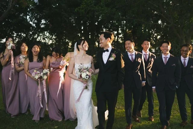 A wedding party of eight, including the bride in a white gown holding a bouquet, the groom in a black tuxedo, and six bridesmaids and groomsmen dressed in formal attire, standing outdoors among trees.