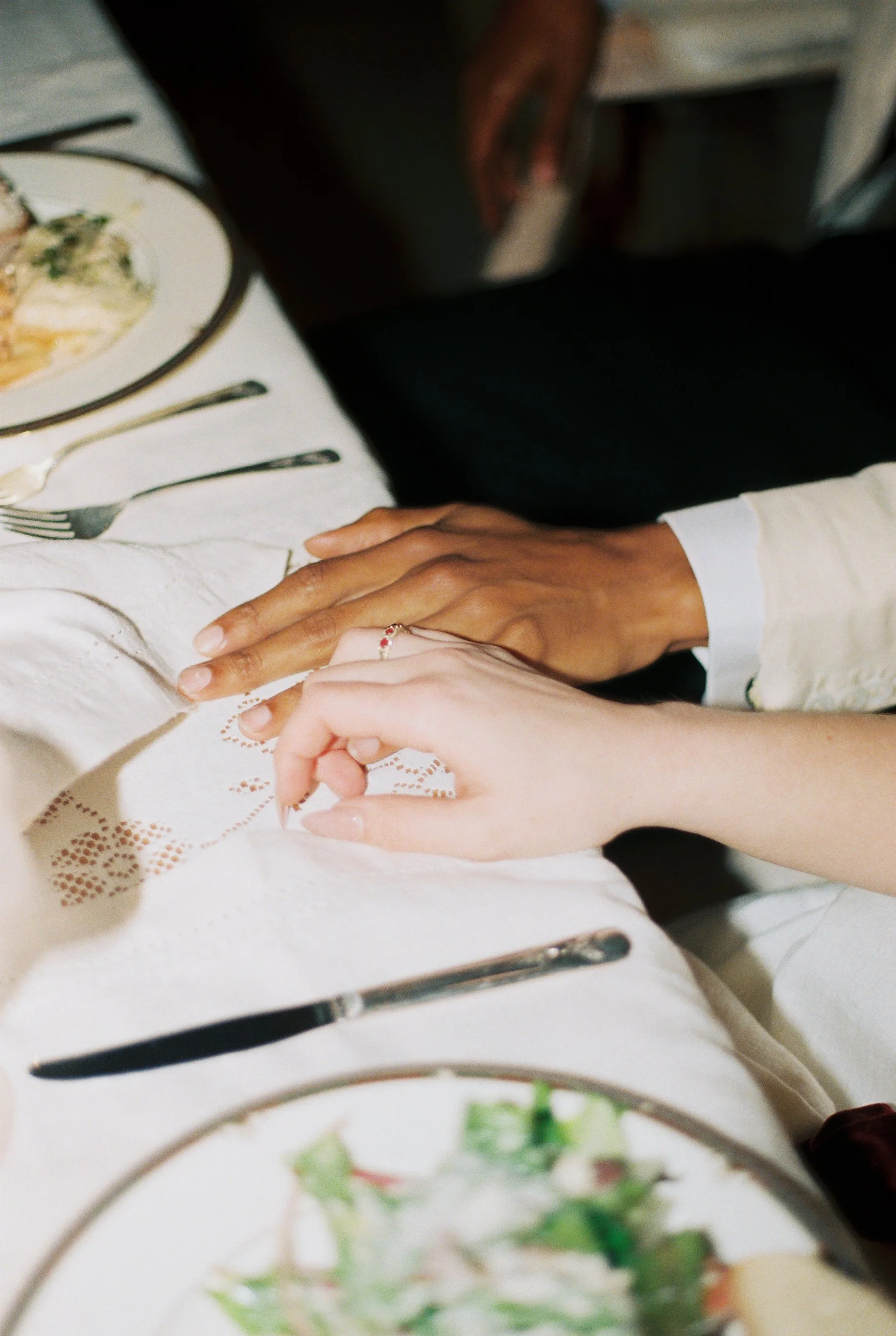 A couple holding hands across a table at a restaurant, with wedding or engagement rings visible. The table is set with dishes and utensils.