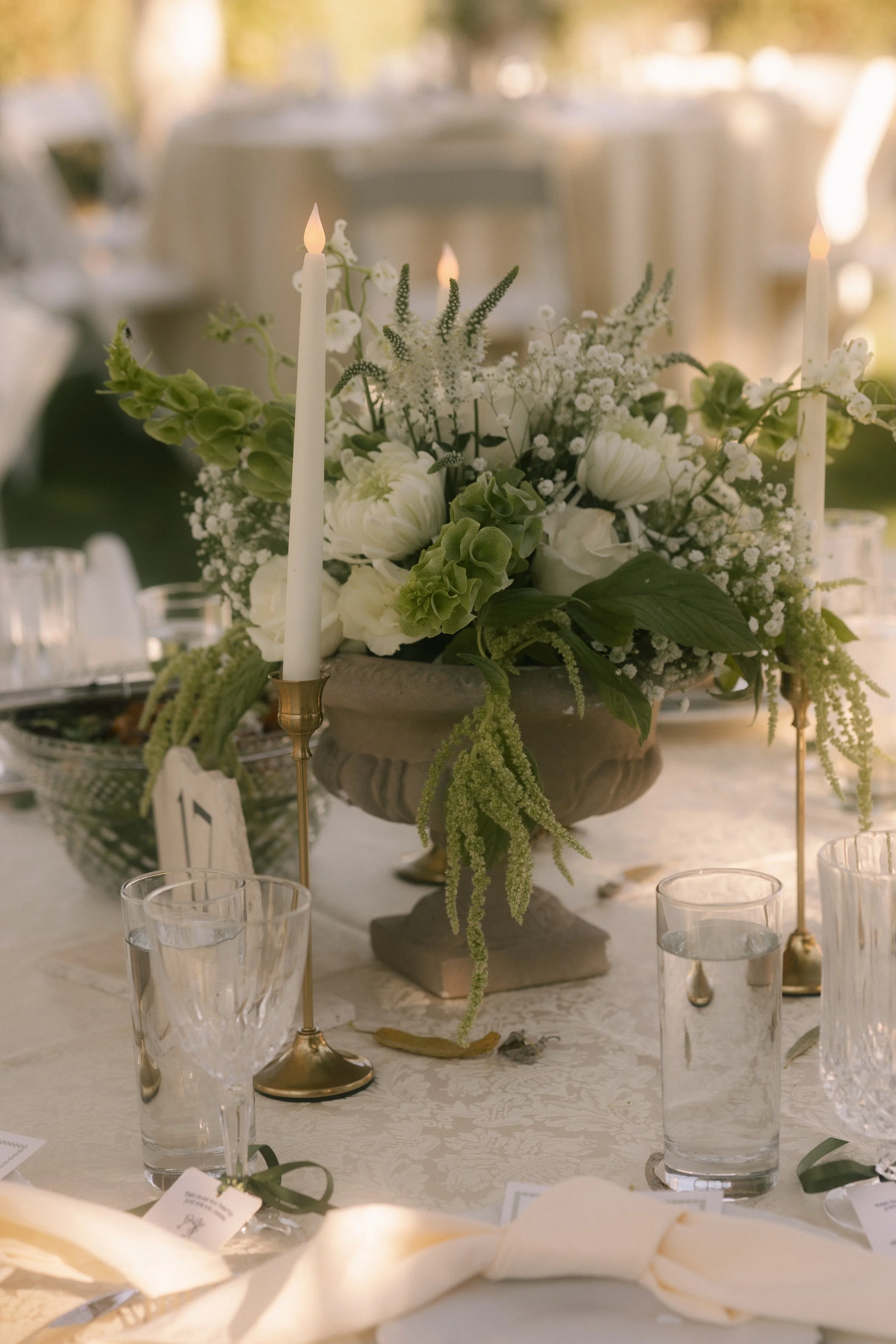 A table centerpiece featuring white flowers in a decorative urn, flanked by tall white candles on gold holders, with glasses and place cards set on a white tablecloth at a formal event.